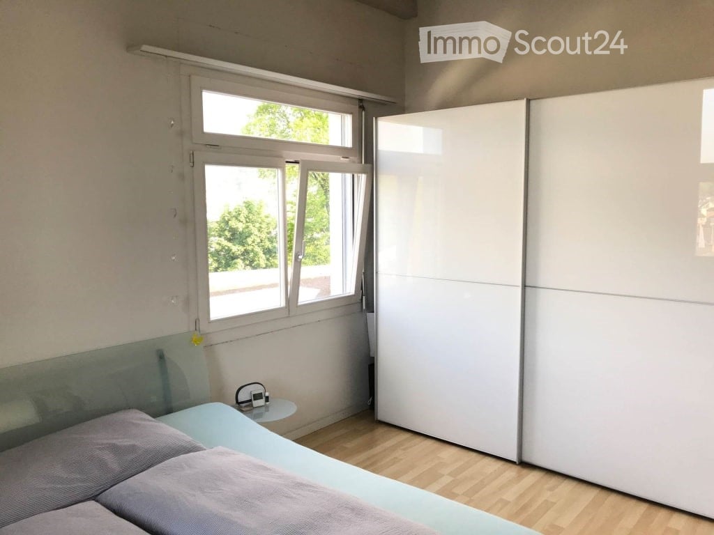 Modern bedroom with a light blue bed, grey bedding, a white wardrobe, and a small nightstand with a lamp and speaker.