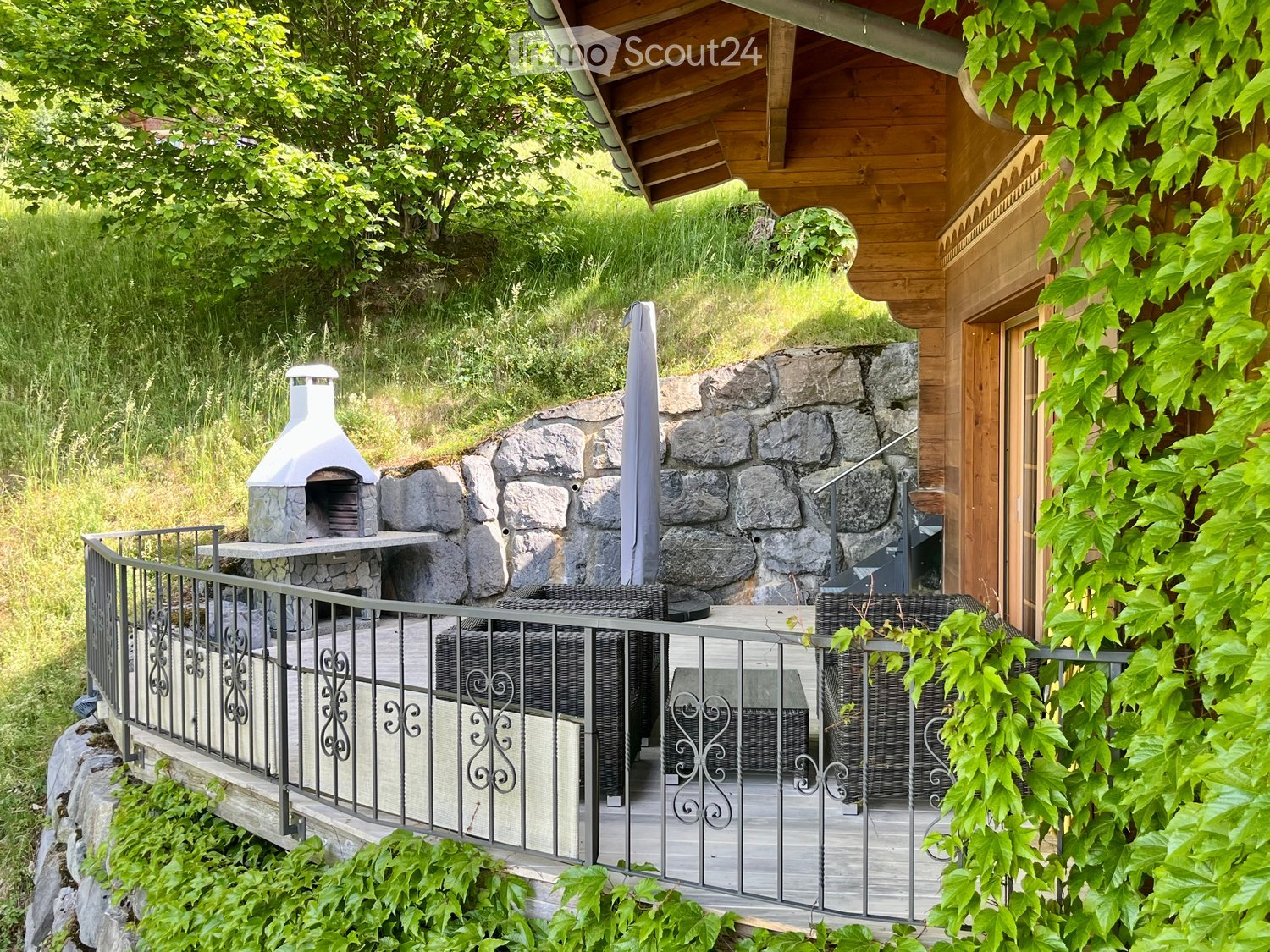 Balcony with a stone pizza oven, wicker chairs, and a metal railing, surrounded by greenery and a wooden house.