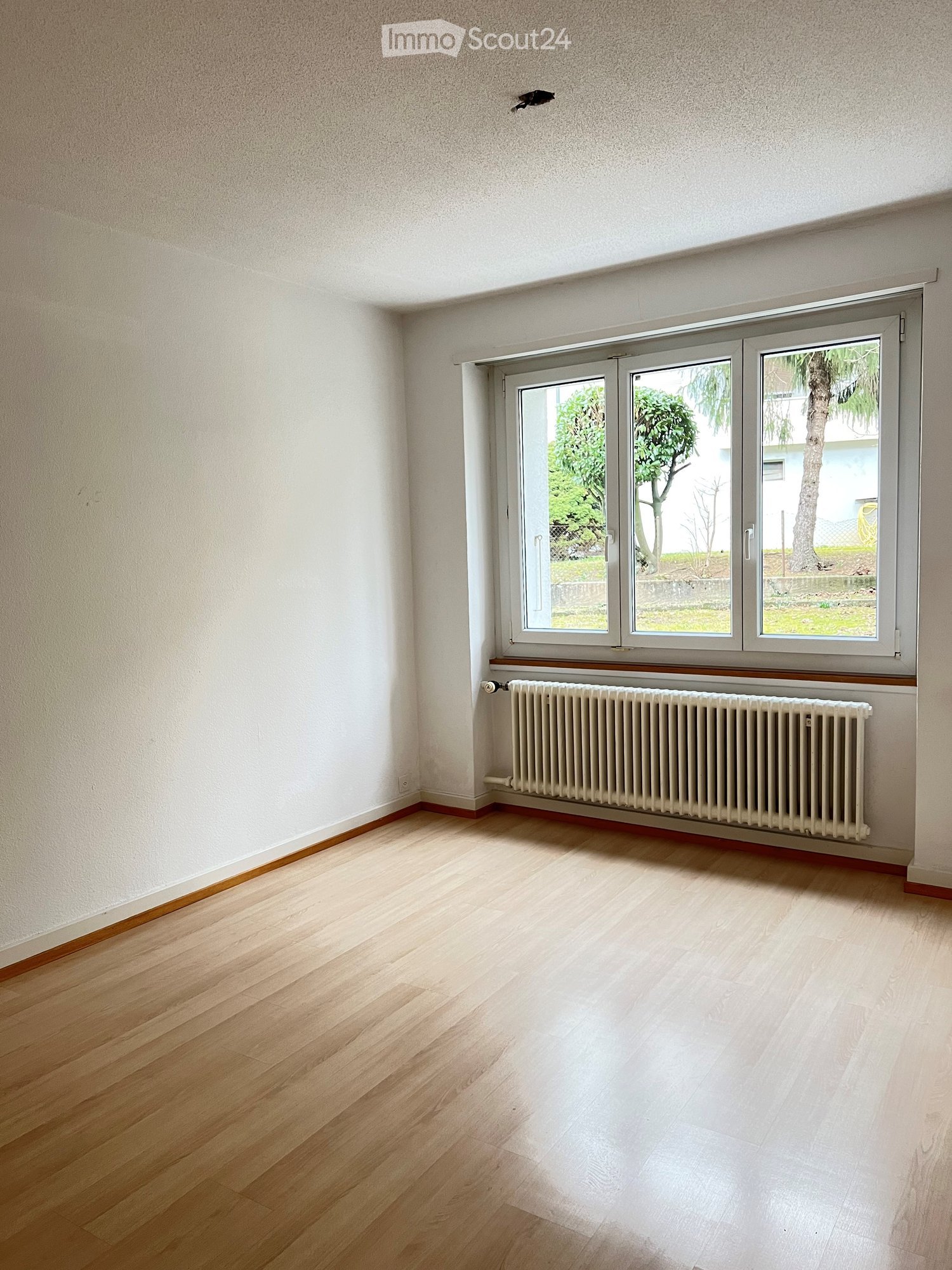 Empty room with light wooden flooring, white walls, and a large window with a radiator below it.