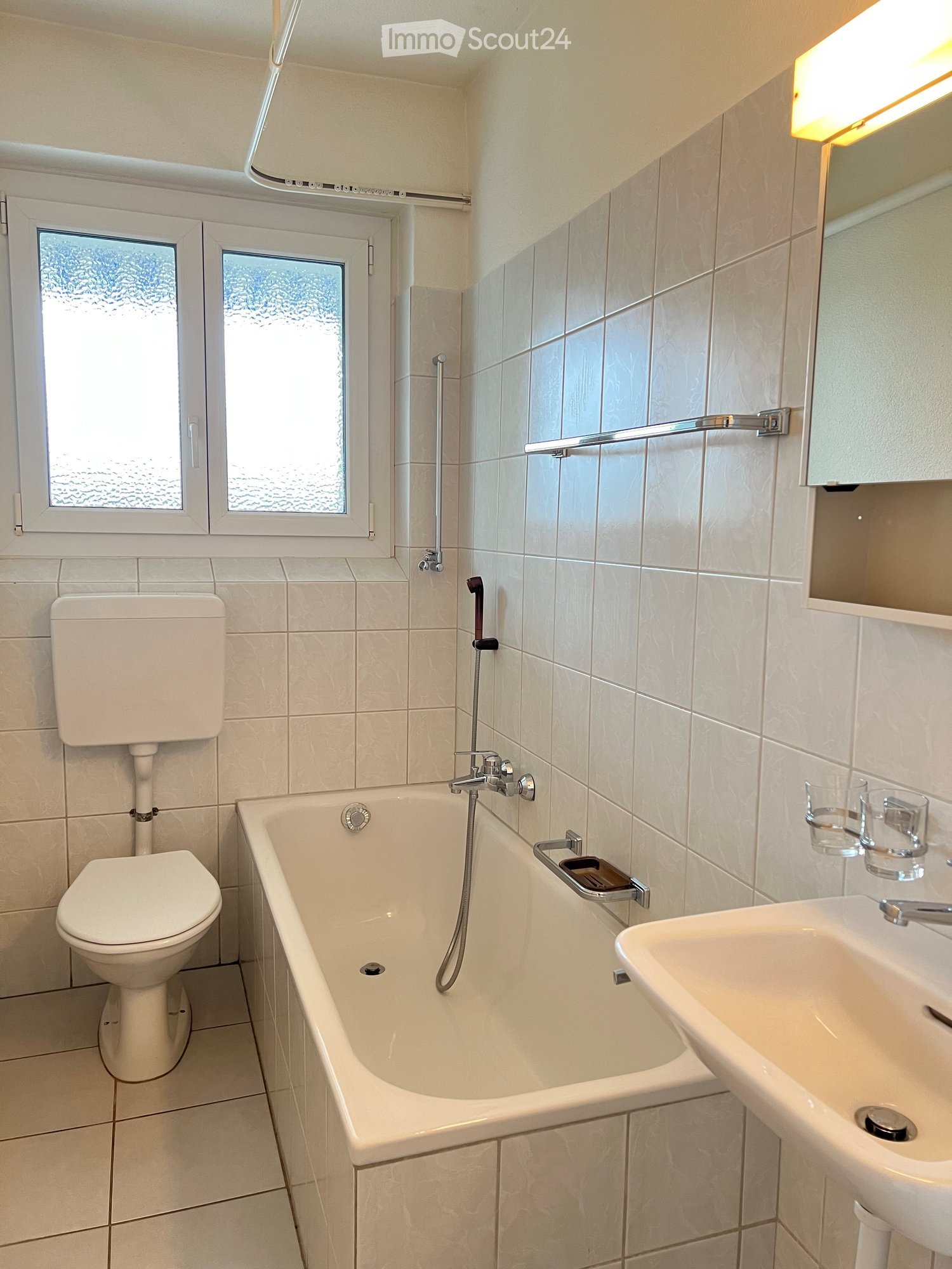 Modern bathroom with white tiles, a bathtub, toilet, sink, and a mirror above the sink. The room features a window with frosted glass and a built-in shelf with cups.
