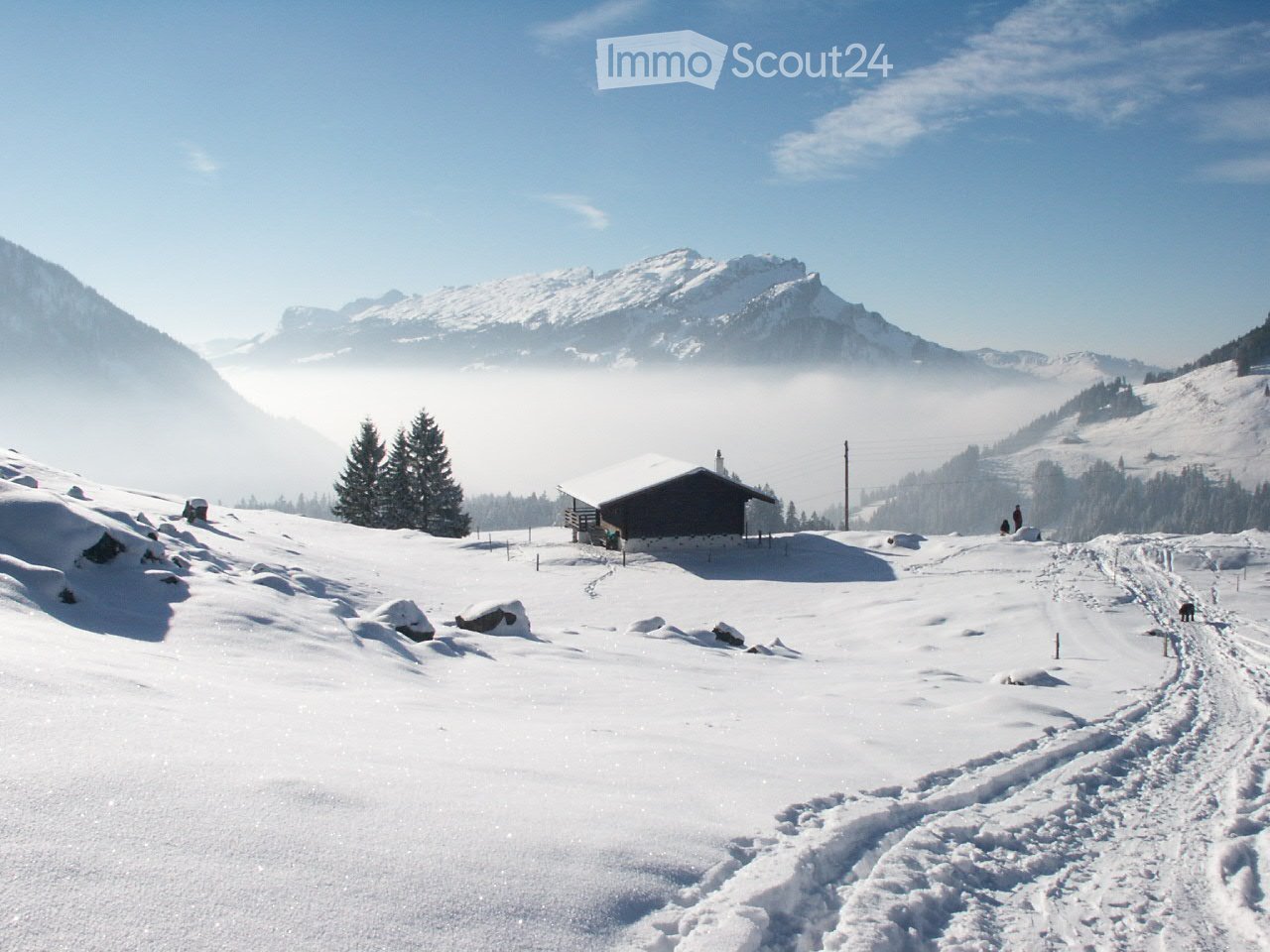 A snow-covered mountain landscape with a small wooden cabin, surrounded by snow-covered trees and mountains under a clear blue sky.