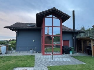 Modern two-story house with dark blue exterior, large red-framed glass entrance, and a small patio area with a grill and outdoor furniture.