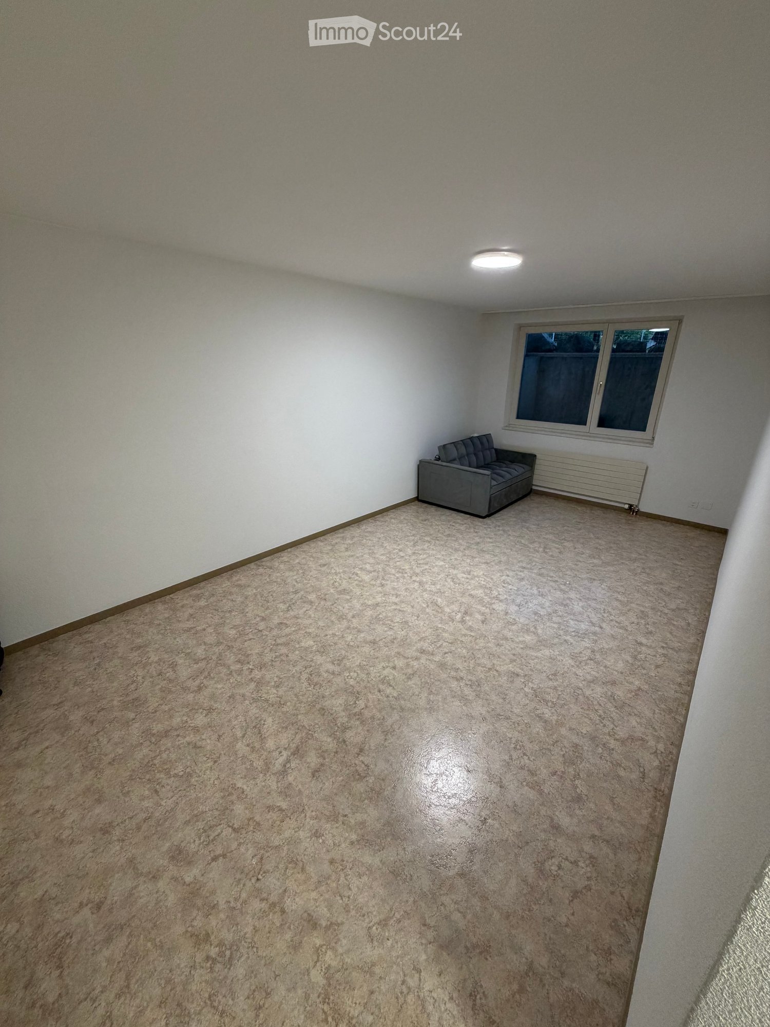 A small, carpeted room with white walls and a single window. Contains a grey sofa against the wall.