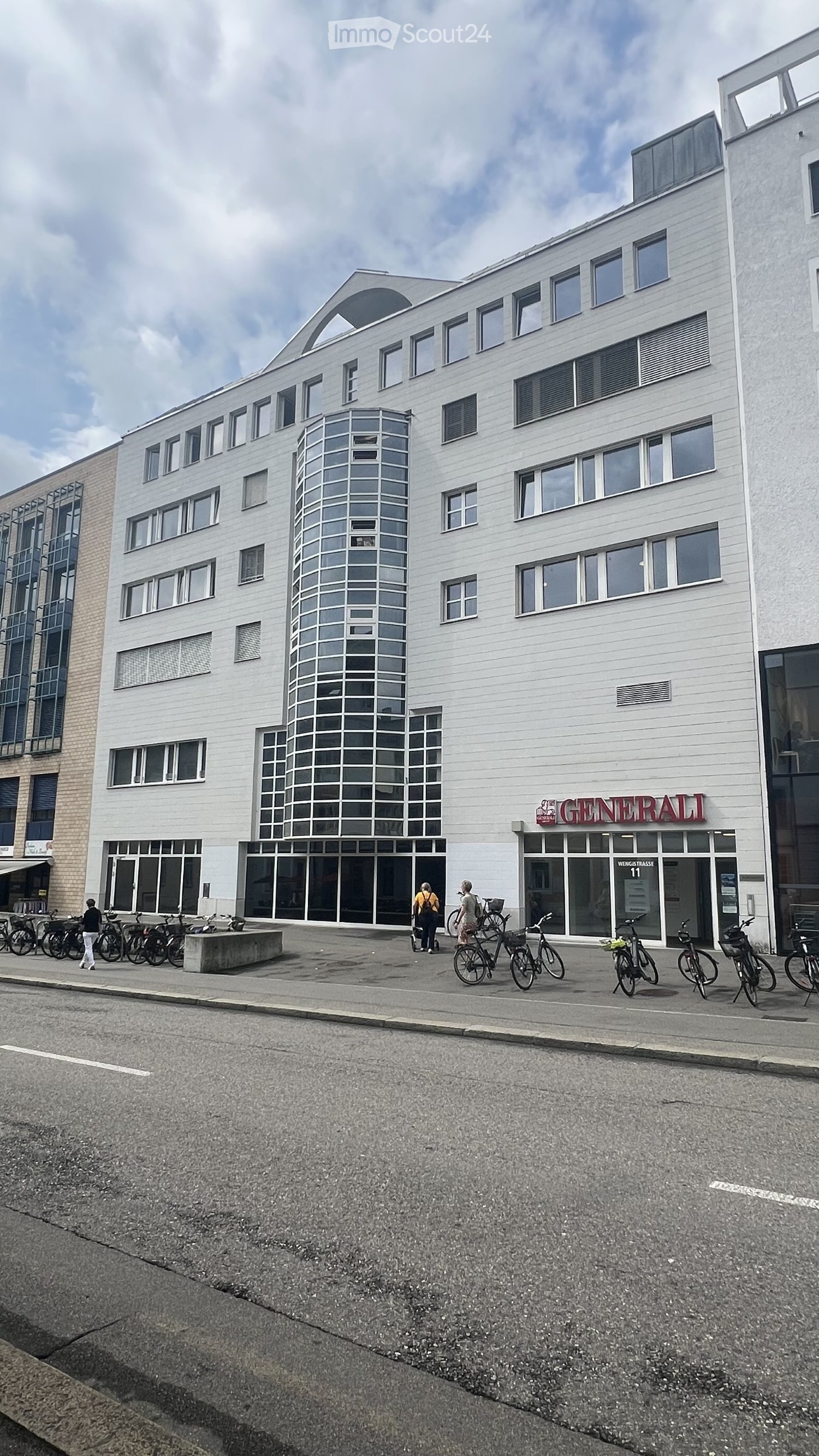 Modern multi-story building with white facade, large windows, and a cylindrical tower section. The ground floor has glass storefronts with signage reading "GENERALI" and "WEIGTIKLASSE 11". Bicycles are parked along the sidewalk in front of the building.