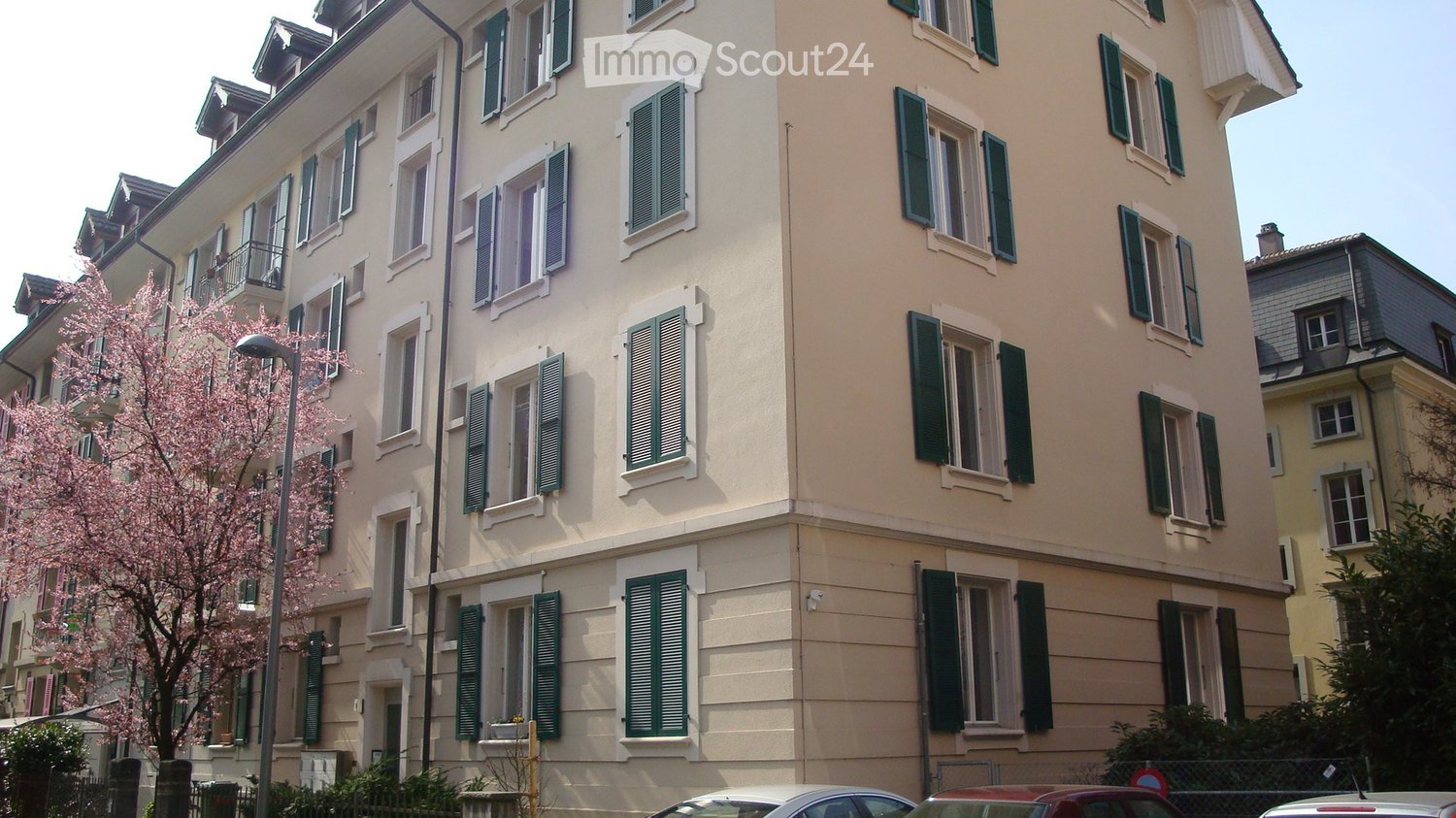A multi-story residential building with light beige walls, green shutters, and multiple windows. The building has a classic architectural style with a flat roof and decorative details.