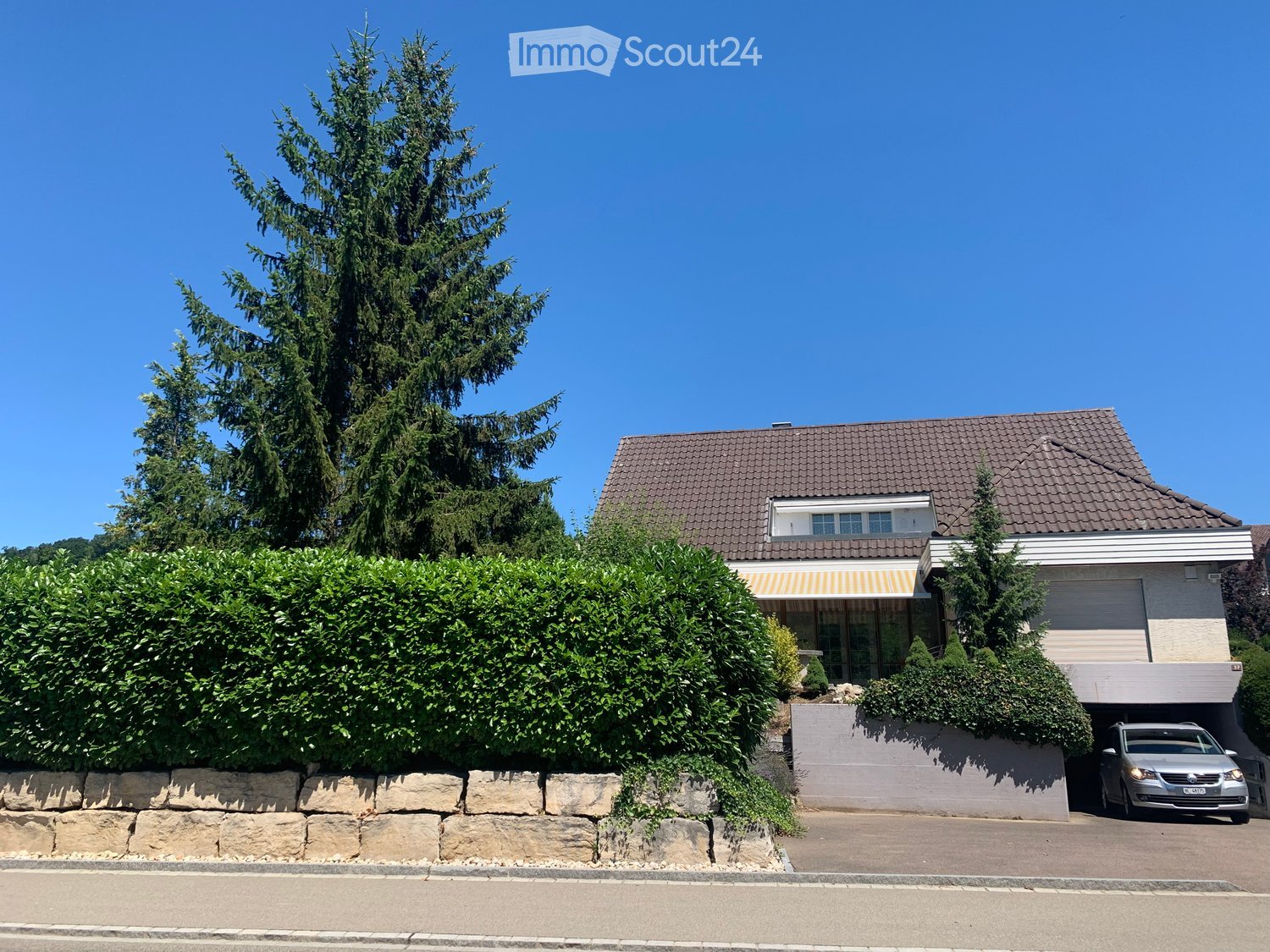 Two-story house with a tiled roof, garage, and surrounding greenery including a large tree and shrubs.