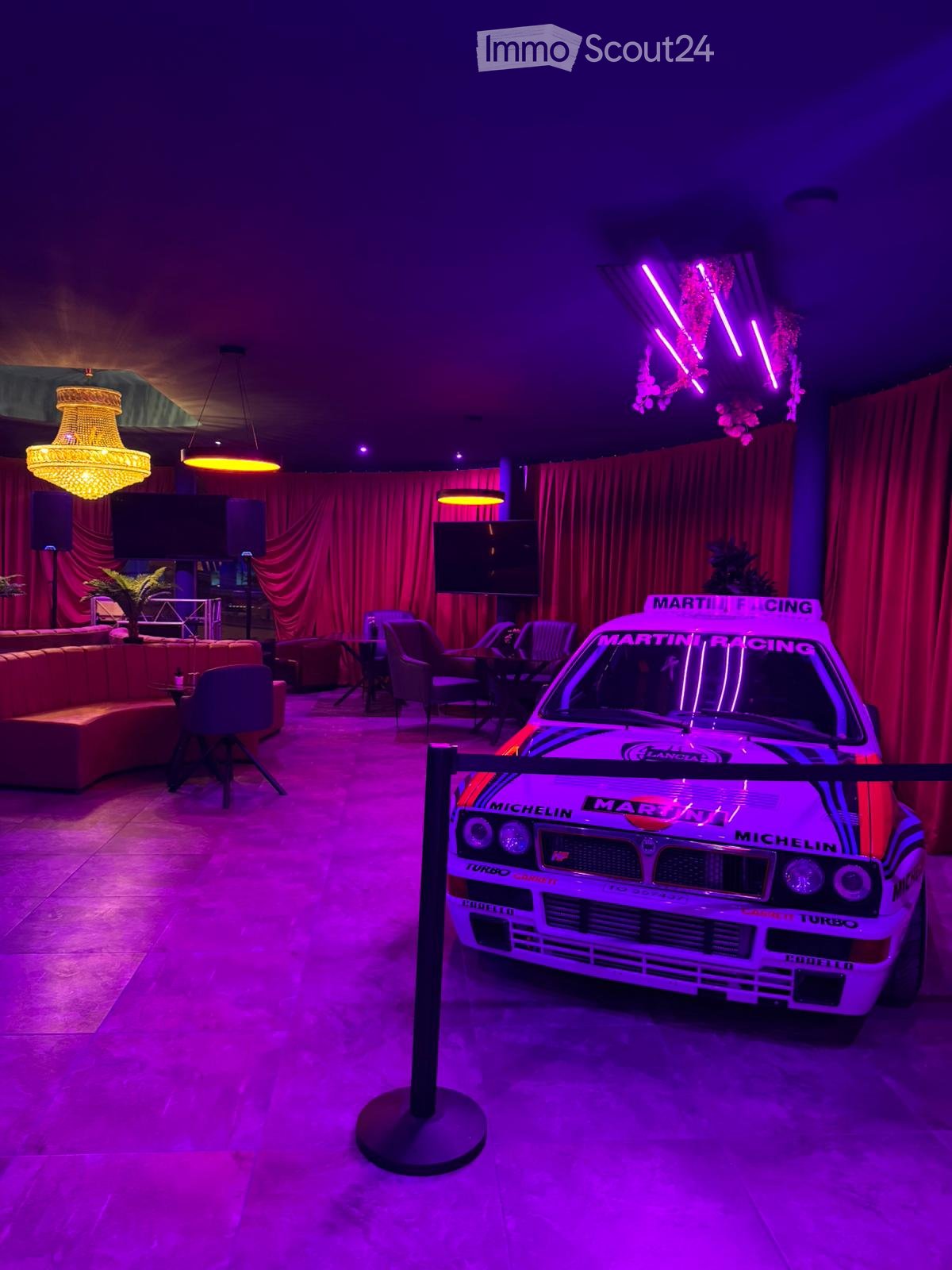 A stylishly furnished room with a vintage racing car prominently displayed, featuring a mix of seating areas, a television, and decorative lighting.