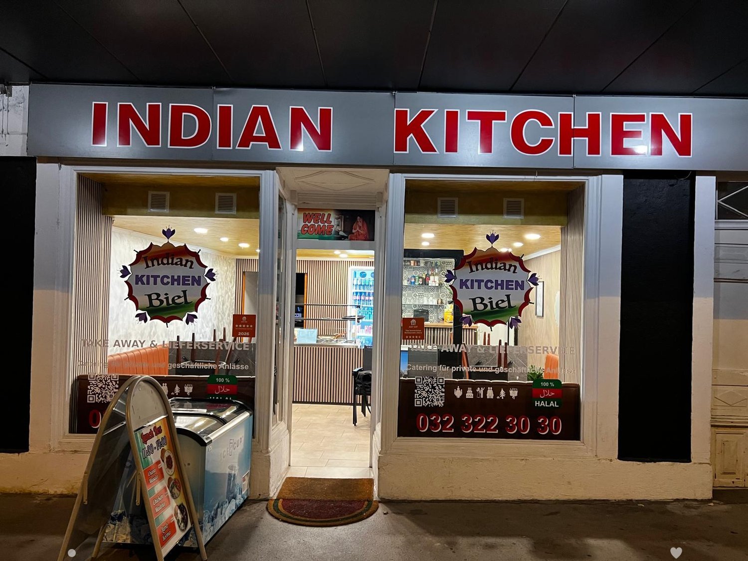 Exterior view of "Indian Kitchen" restaurant with signage, open doorway, and visible interior seating area with tables and chairs.