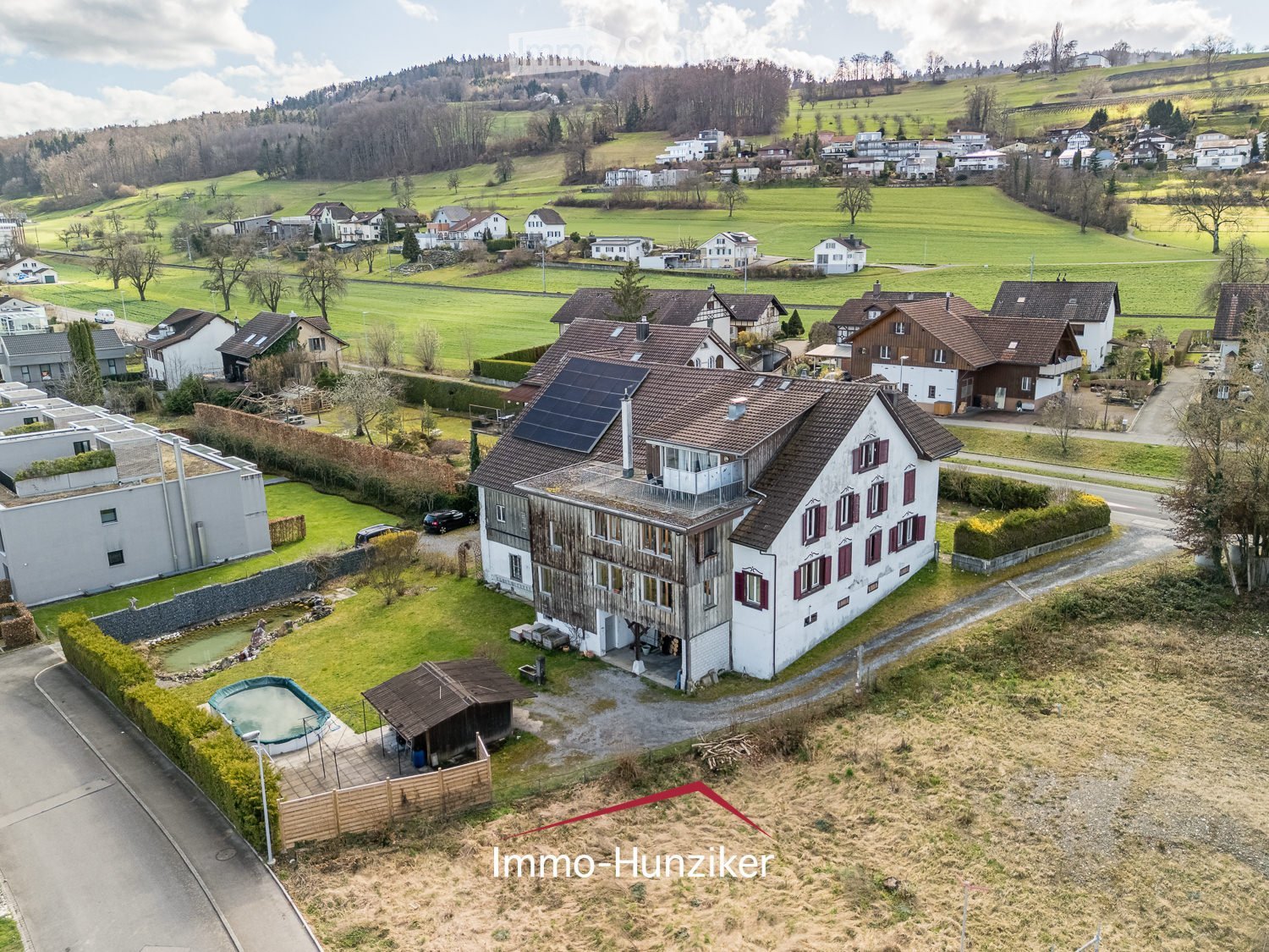 Aerial view of a residential house with a swimming pool, surrounded by greenery and neighboring houses in a rural setting.