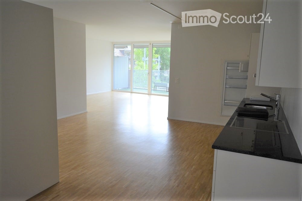 Empty room with wooden flooring, white walls, and a small kitchen area with a sink and cabinets. Large sliding glass doors lead to a balcony.