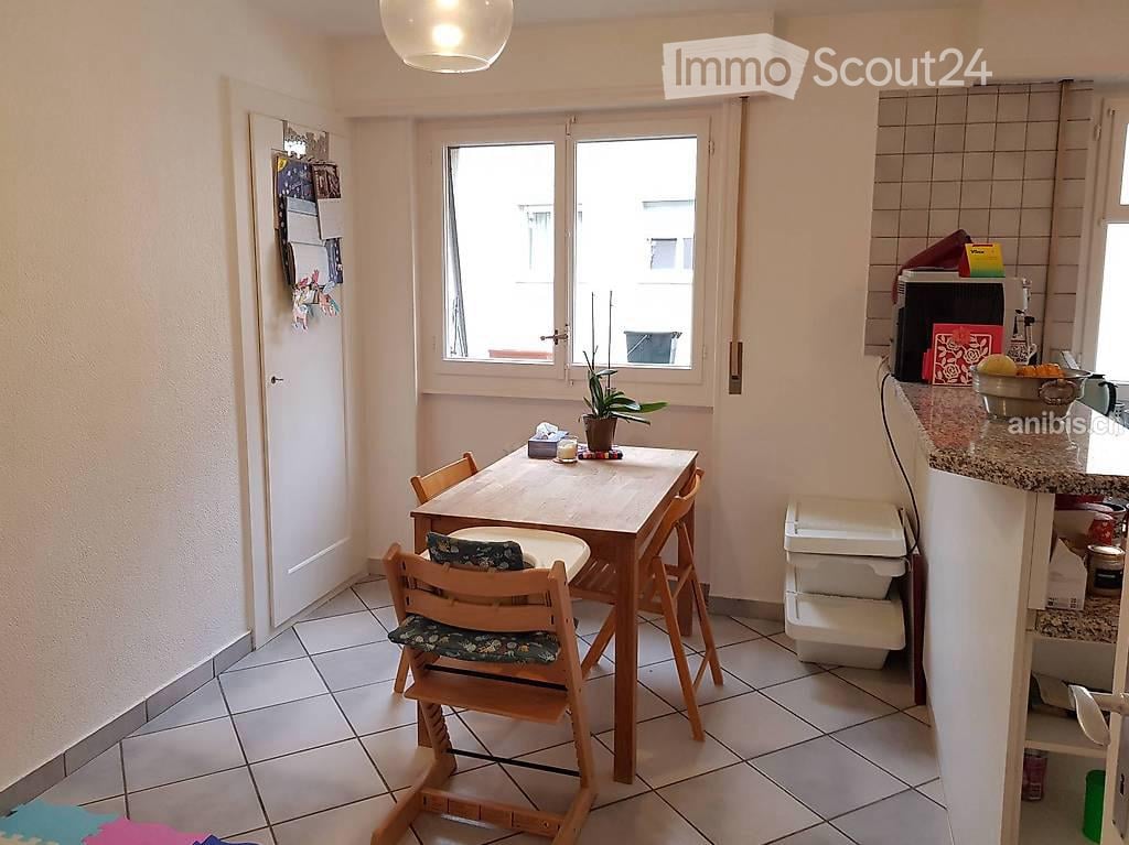 A small, well-lit dining area with a wooden dining table and chairs, a potted plant on the table, and a microwave on the counter.