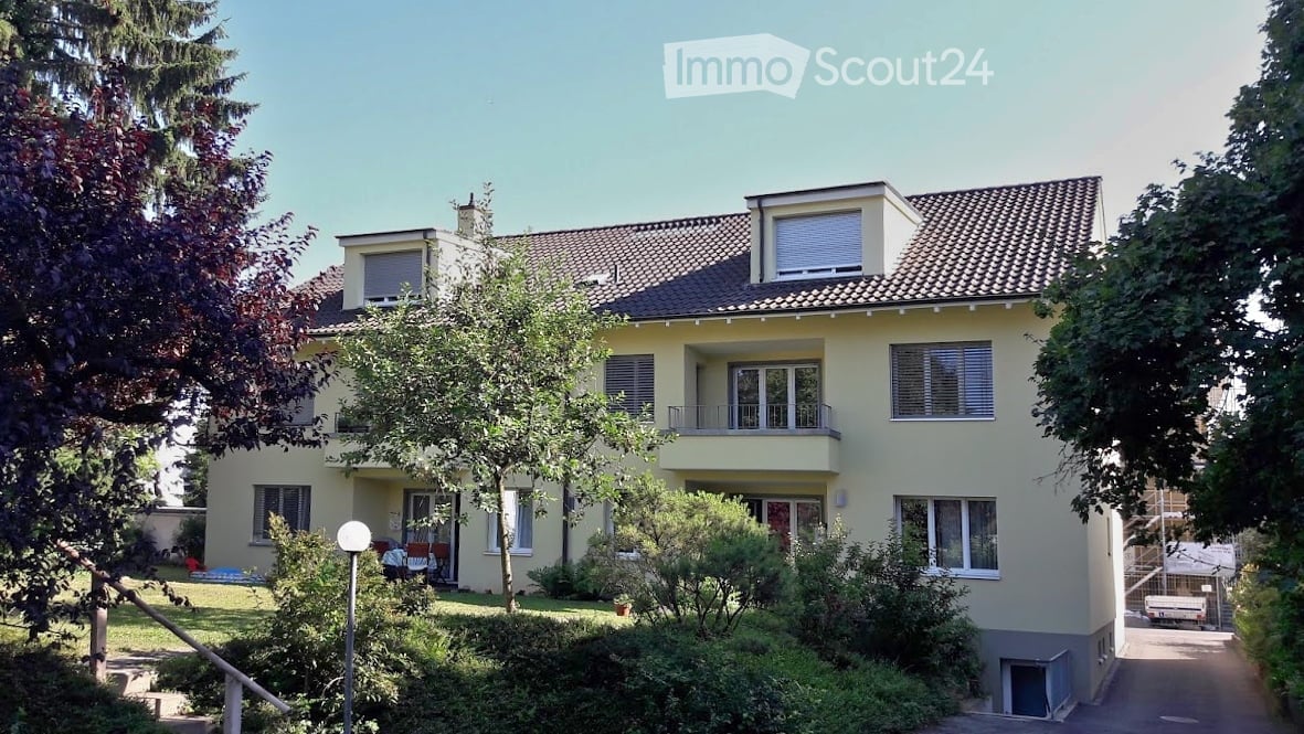 Two-story house with yellow exterior, multiple windows, and a balcony on the upper floor. The house is surrounded by greenery and trees.