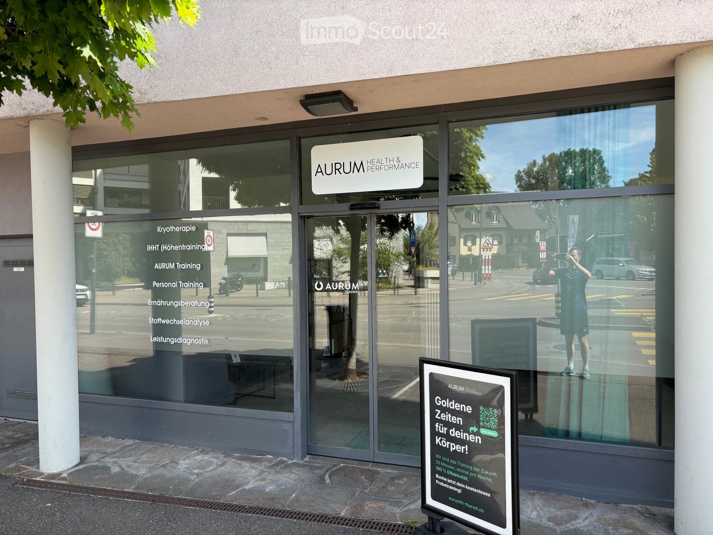 The image shows the exterior of a modern building with large glass windows and a sign reading "AURUM HEALTH & PERFORMANCE". A promotional sign in front advertises a training program. The building has a clean, contemporary design with a visible entrance an
