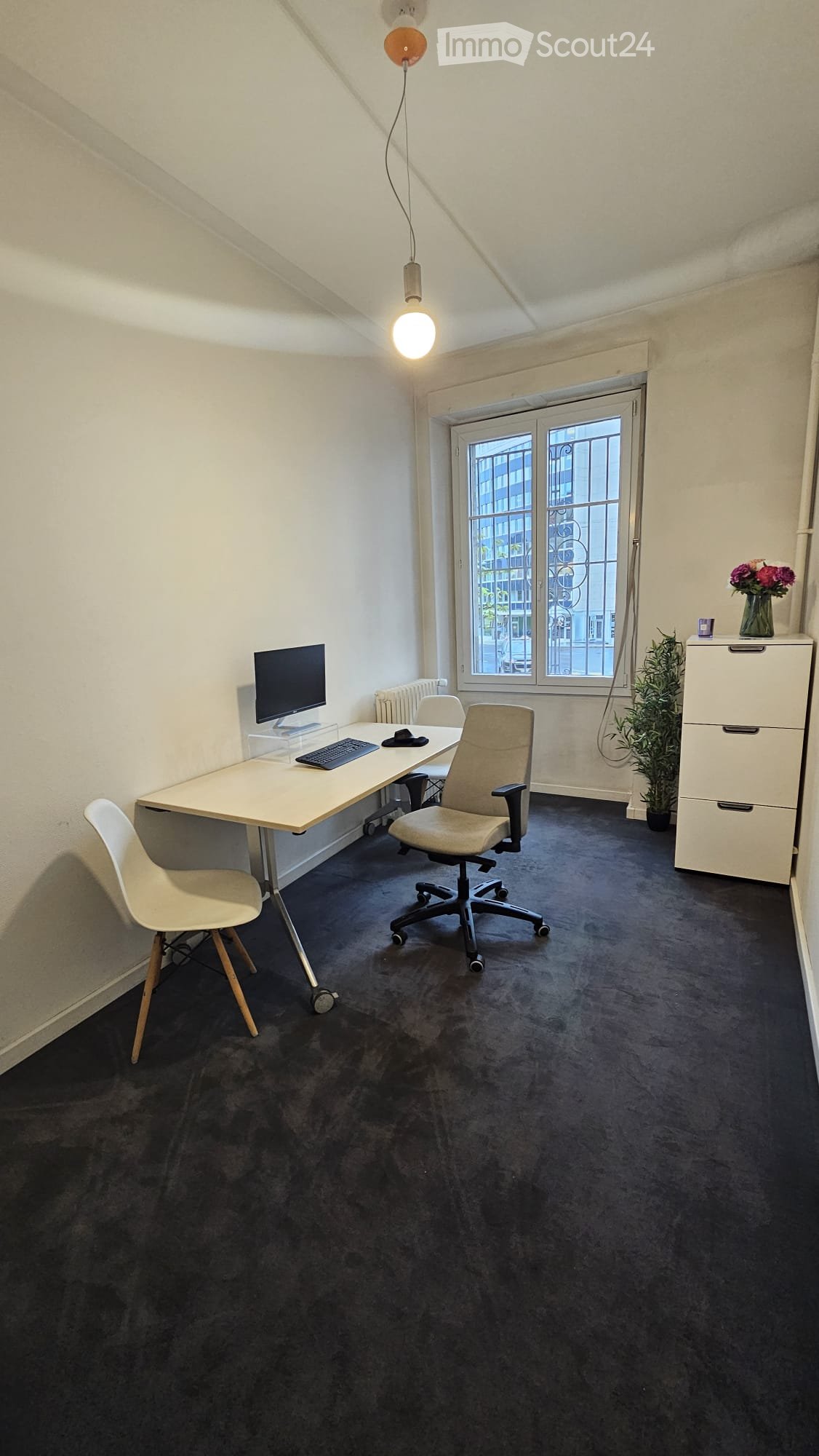 A modern, furnished room with a desk, computer, chairs, filing cabinet, and decorative plant. The room has a large window with a view of the street and is illuminated by a hanging light fixture.