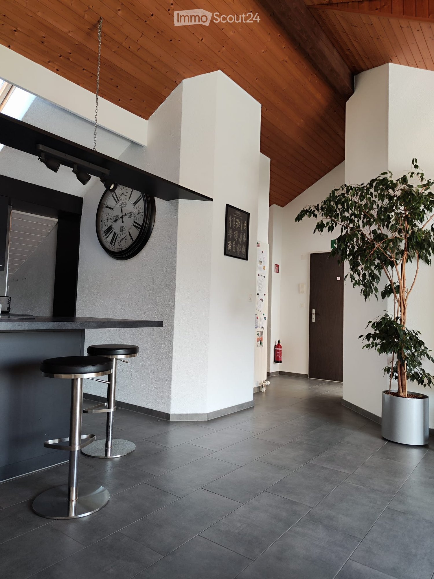 Modern hallway with grey tiled floor, white walls, wooden slanted ceiling, two black stools, a large clock, a framed picture, and a potted plant.
