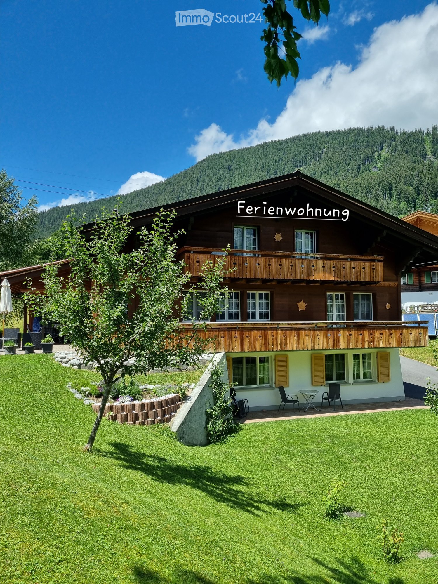 A two-story chalet with a balcony, surrounded by a well-maintained garden and a mountain backdrop.