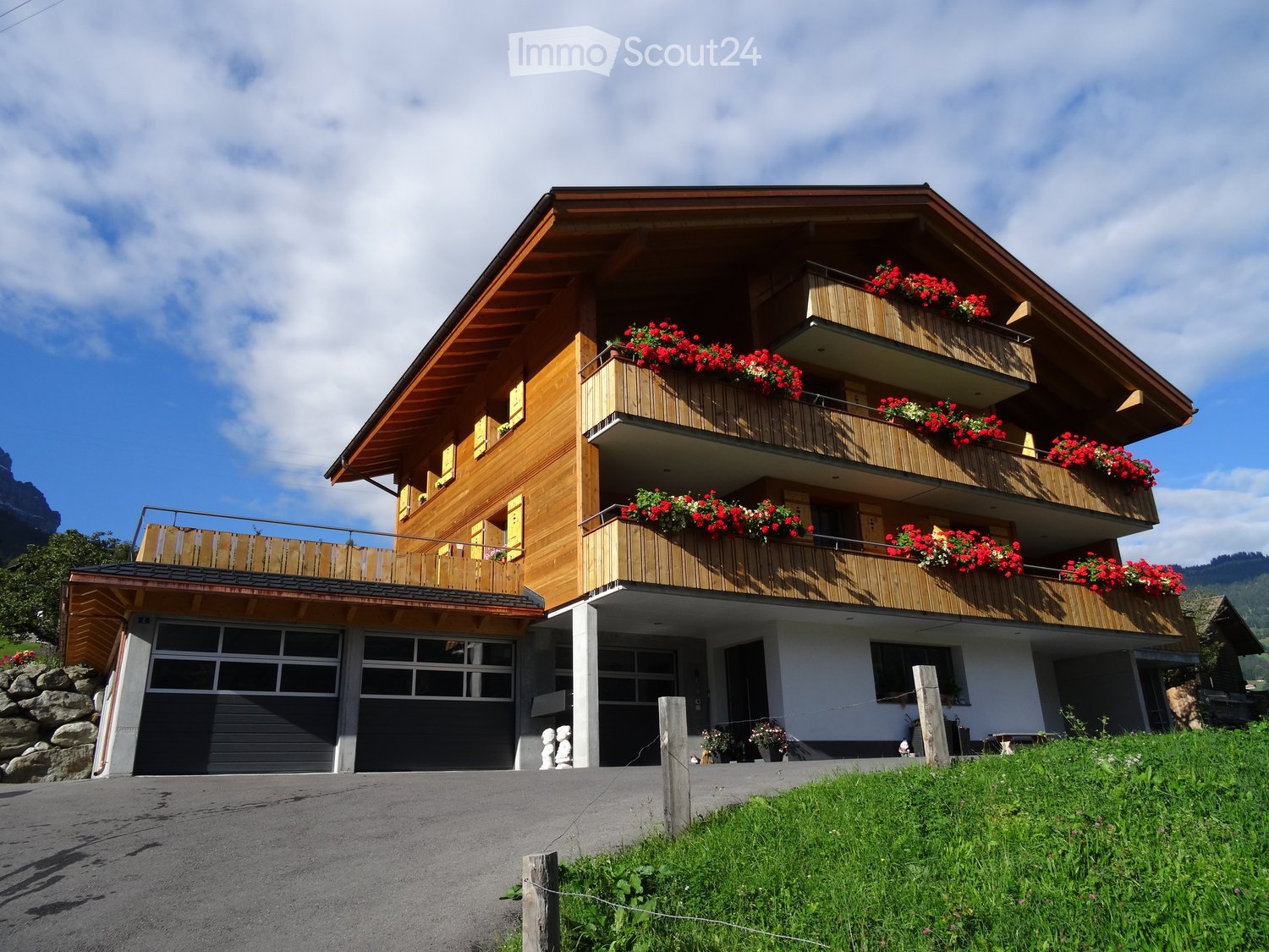 A three-story wooden house with balconies adorned with red and white flowers, a garage, and a stone foundation.