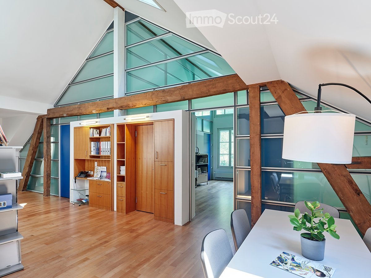 Bright, modern room with wooden flooring, large glass windows, and exposed wooden beams. Features a white table with a potted plant, chairs, a wooden cabinet with shelves, and a desk area with a telephone and books.