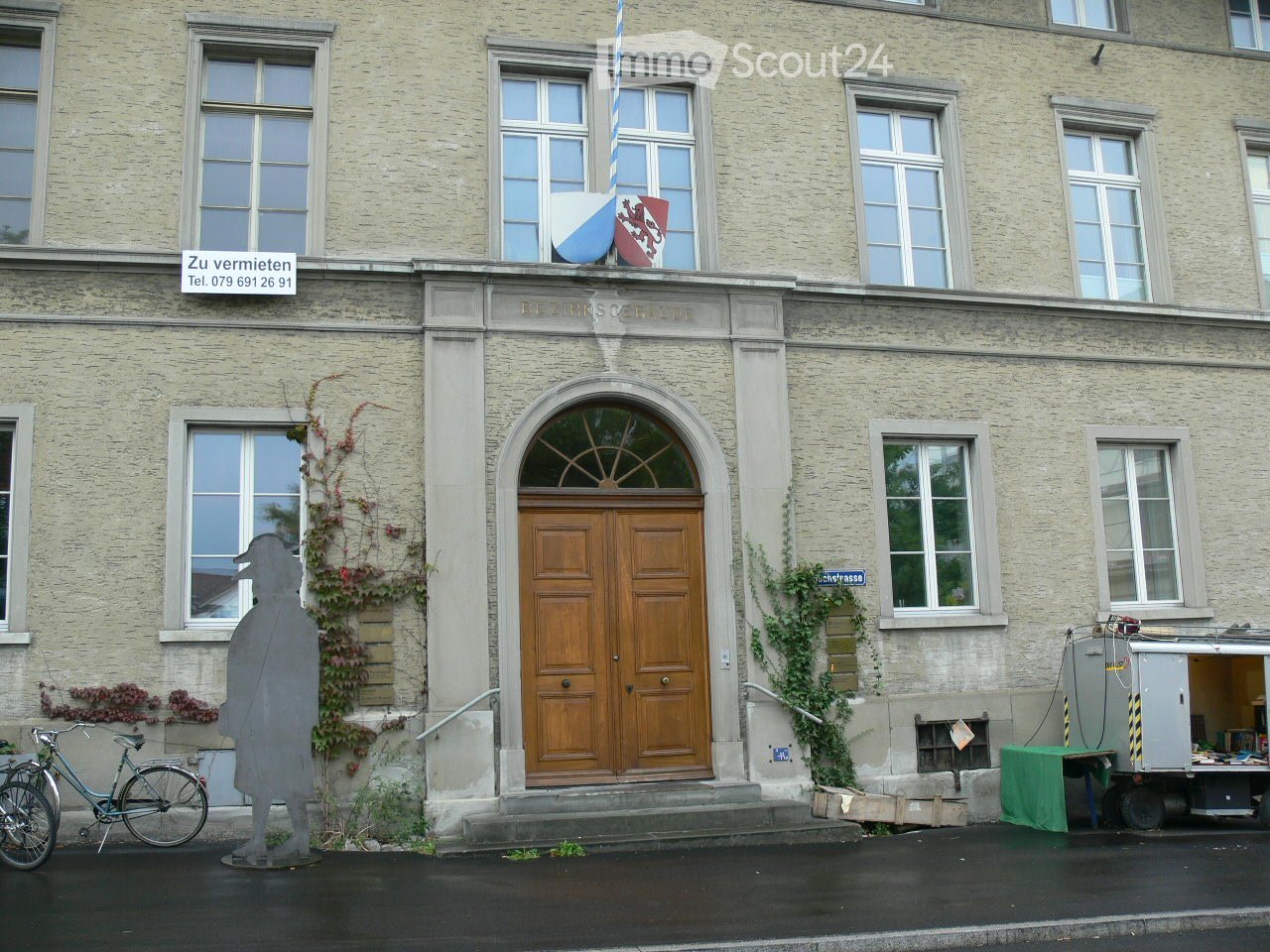 Facade of a multi-story building with large windows, a central wooden double door, and a flagpole with a flag and coat of arms above the entrance.