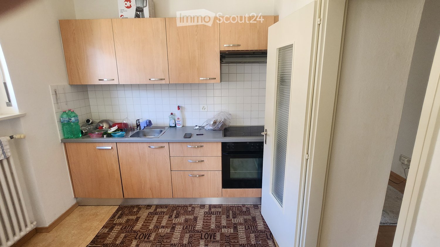 The kitchen features light wooden cabinets, a stainless steel sink, and a built-in oven. The countertop has various items including bottles, a blender, and cleaning supplies.