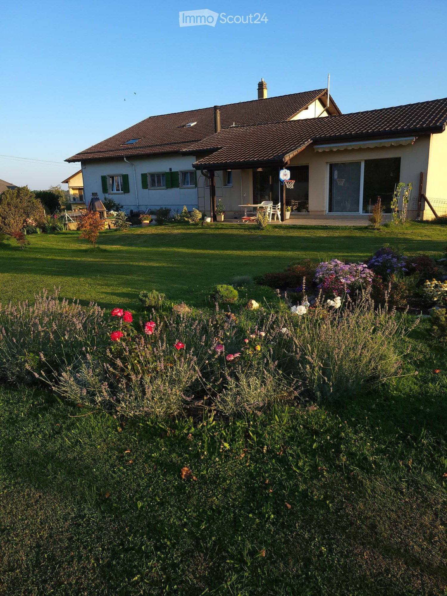 A well-maintained garden with a variety of colorful flowers and shrubs, leading to a single-story house with a tiled roof and green shutters. The house features a covered patio area with outdoor furniture and a basketball hoop.