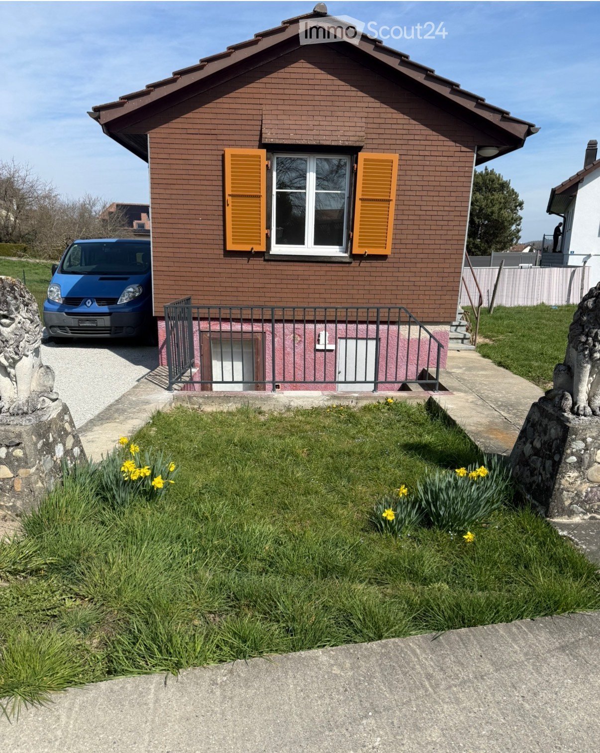 Two-story house with brown brick exterior, white-framed windows with orange shutters, a small garden with yellow flowers, and a blue van parked in the driveway.