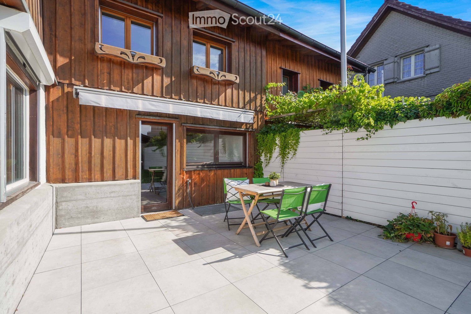 A modern outdoor patio with a tiled floor, a small table with four green chairs, and potted plants along the edge. The patio is adjacent to a wooden house with large windows and a covered entrance.