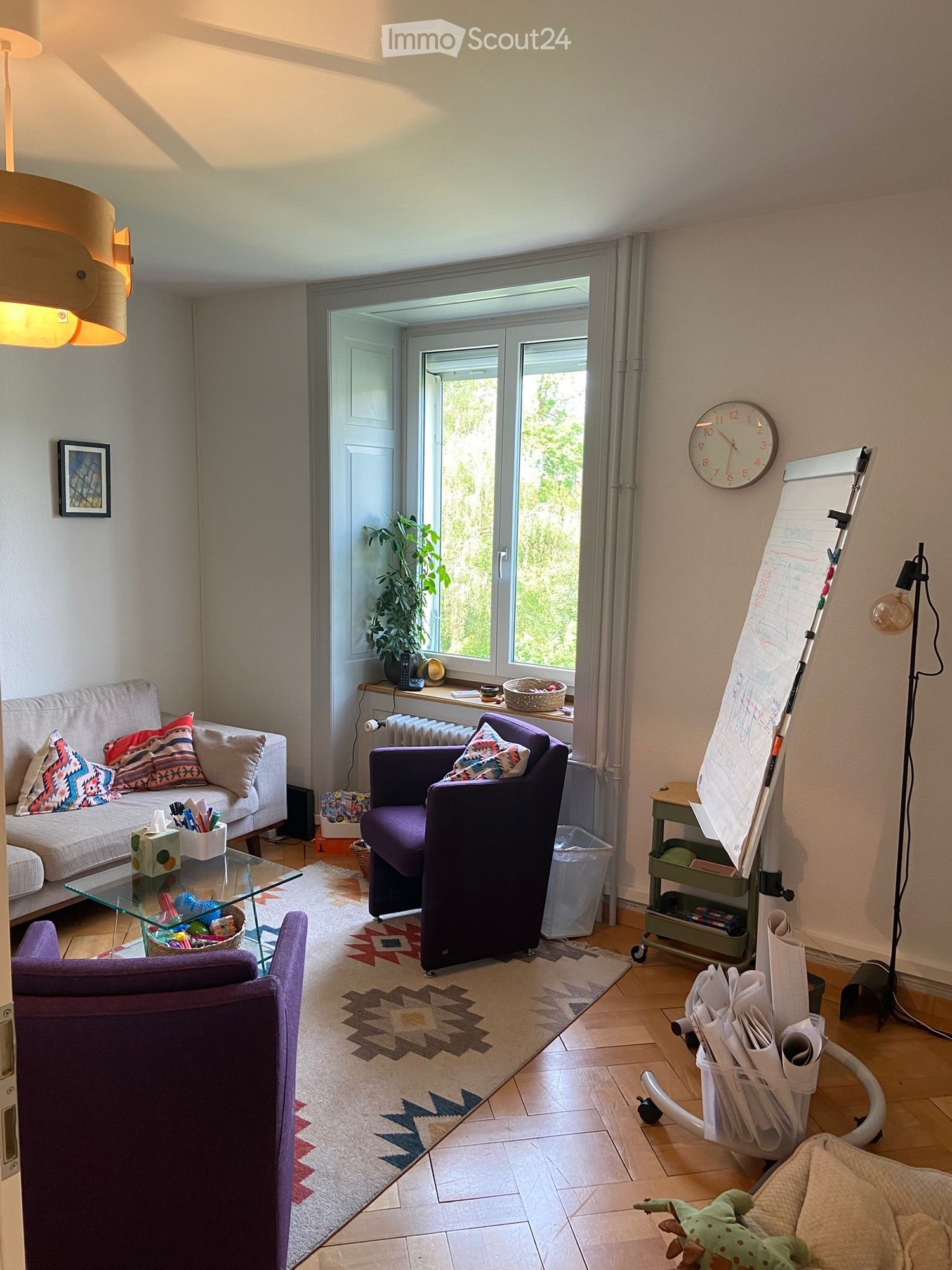 Living room with a sofa, armchair, coffee table, clock, plant, board, lamp, and picture frame. The room has large windows and a view of greenery outside.