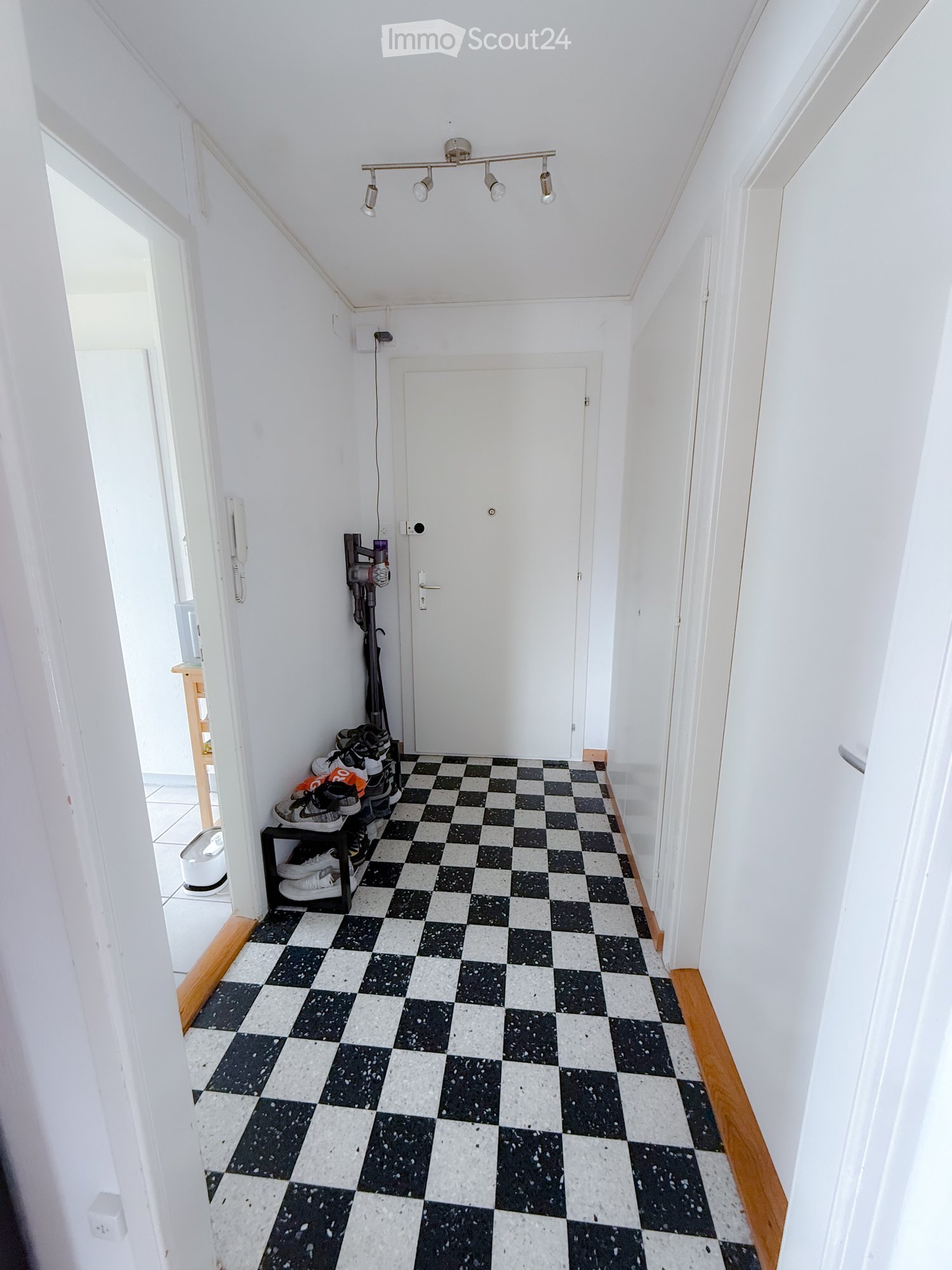 A narrow hallway with white walls, a black and white checkered floor, and a door at the end. A small shelf holds shoes, and a vacuum cleaner is leaning against the wall.