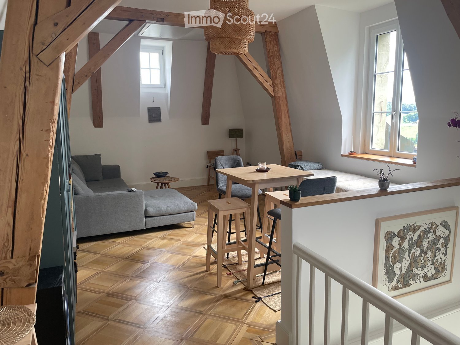 Open-plan living and dining area with wooden flooring, exposed wooden beams, a grey sofa, a coffee table, a dining table with chairs, and a small bed area with a window seat.