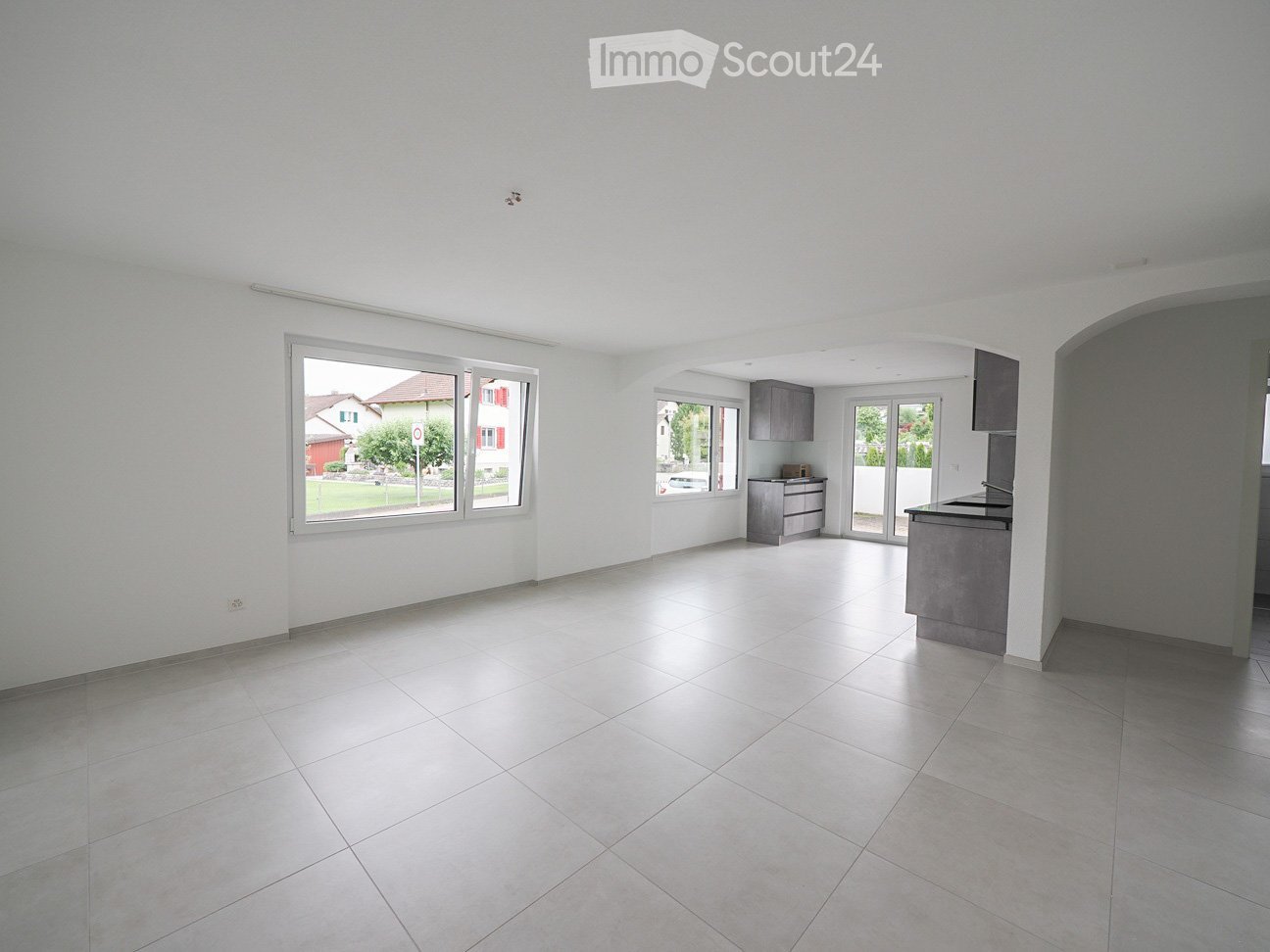 Empty room with tiled floor and white walls, featuring large windows and a small kitchen area with cabinets and a sink.