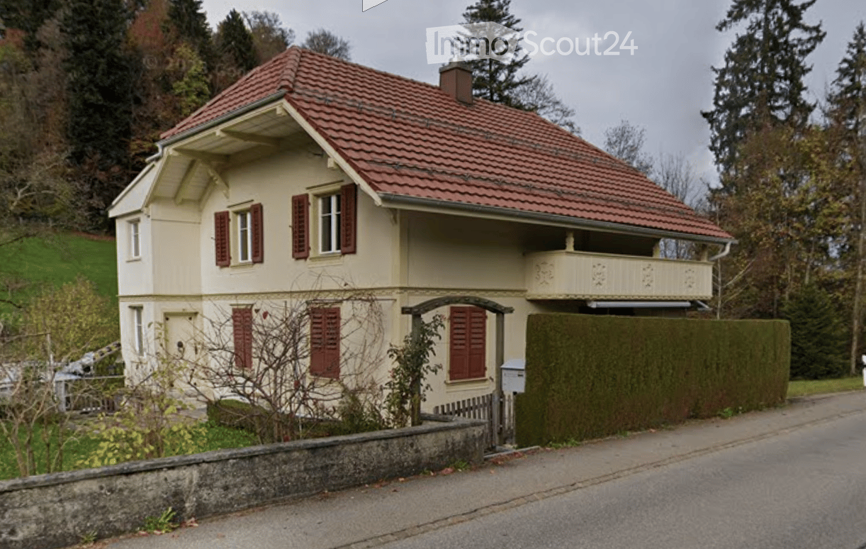 Two-story house with red-tiled roof, cream-colored walls, red shutters, and a small balcony on the upper floor.