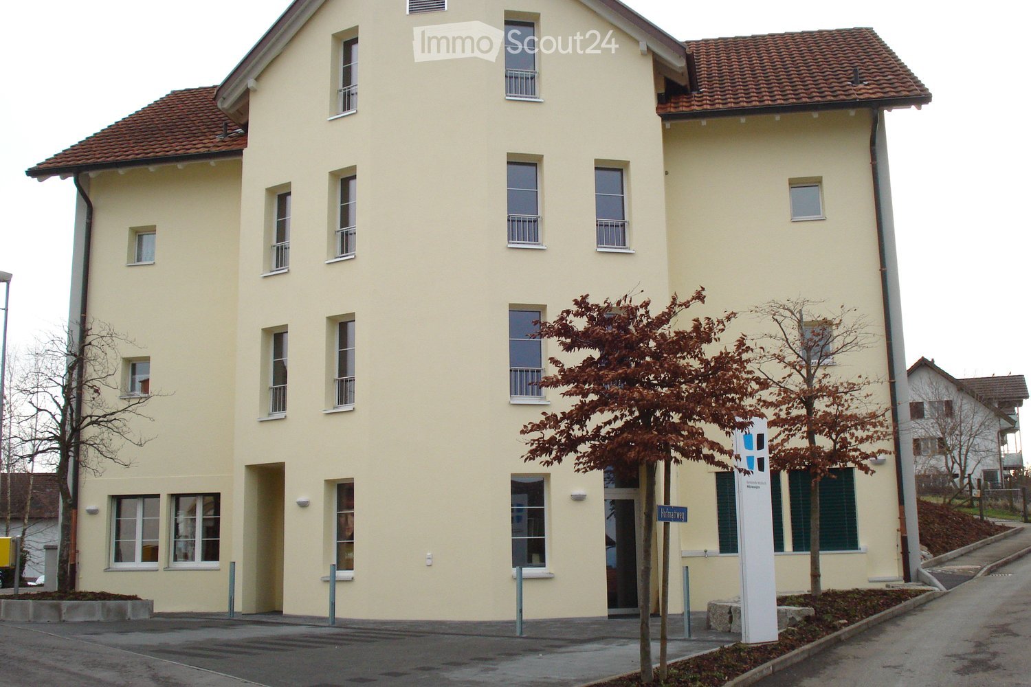 Four-story residential building with light yellow facade, multiple windows, and a red-tiled roof. Features a small tree in front and a street sign.