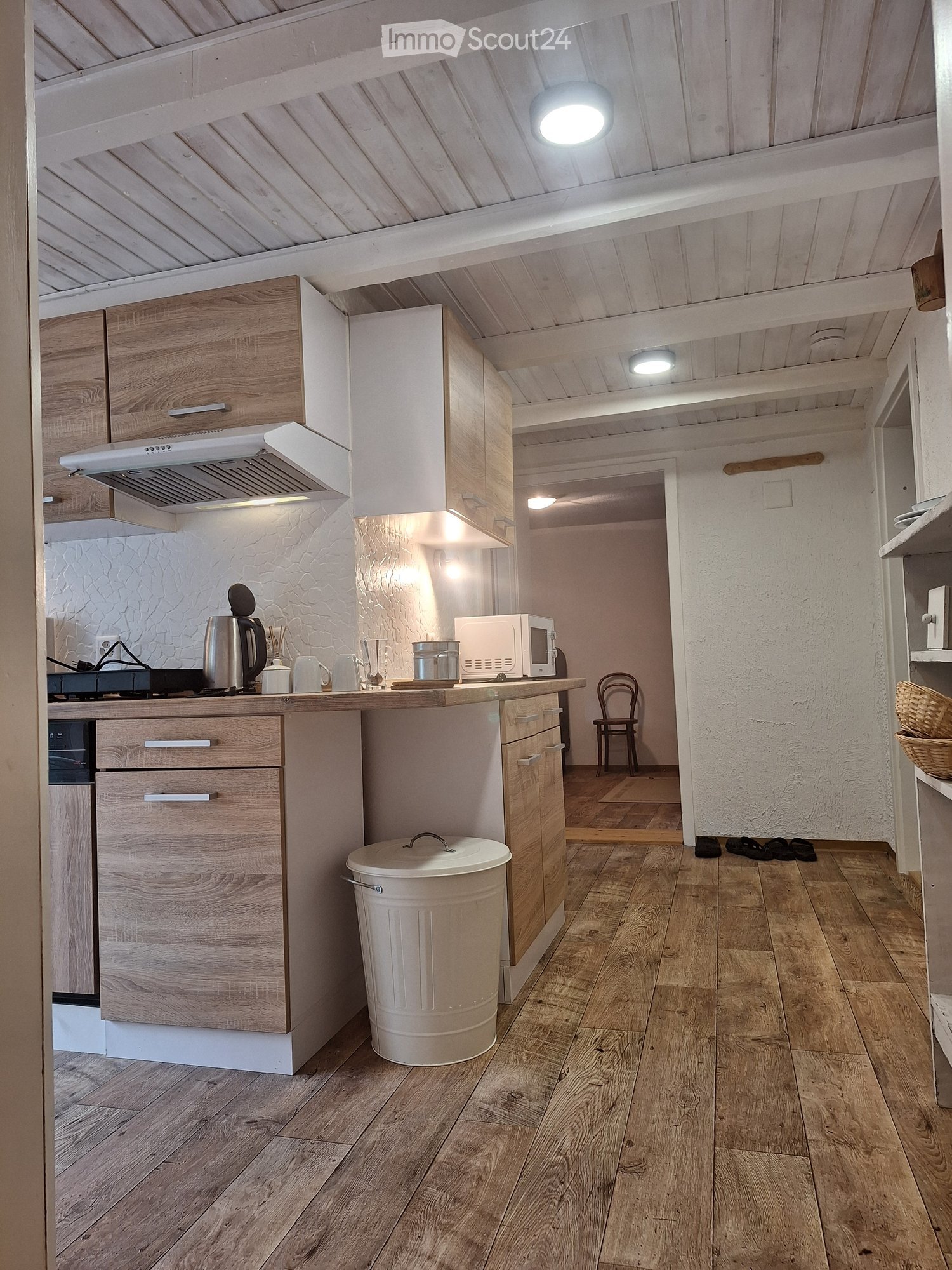 The kitchen features wooden cabinets, a white countertop, and a variety of appliances including a microwave, stove, and oven. The floor is made of light wooden planks, and there is a small chair near the entrance.