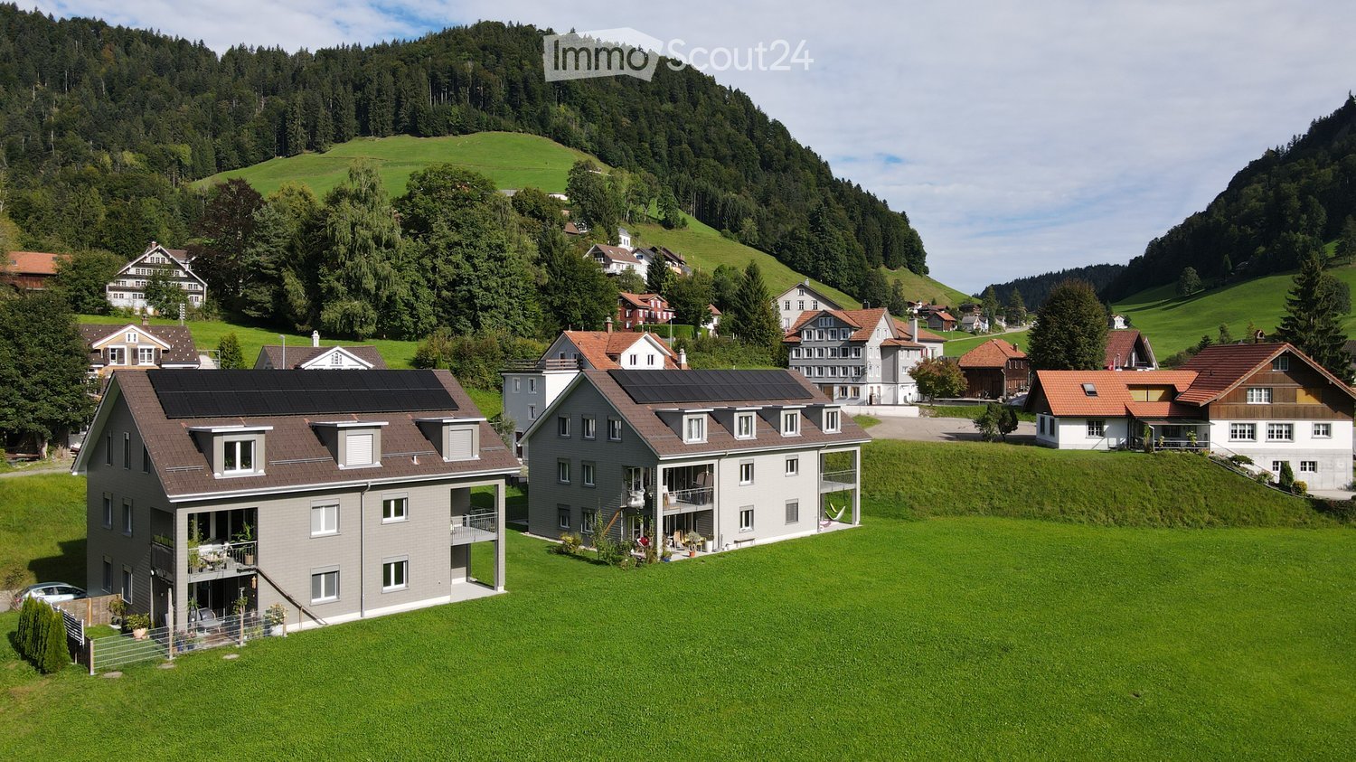 Aerial view of a residential area with multiple houses, surrounded by green hills and trees. Houses have various architectural styles, some with solar panels on the roofs.