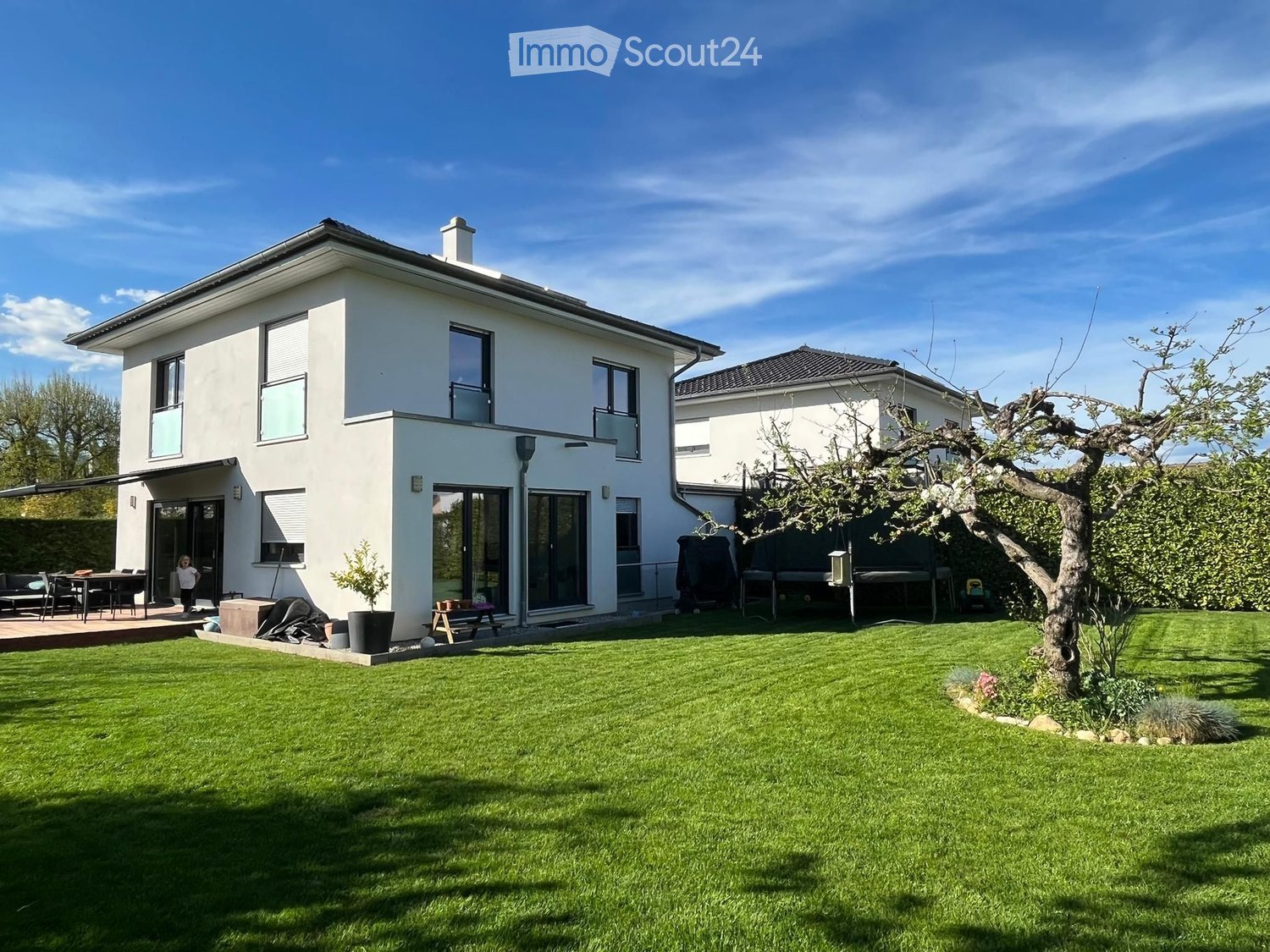 Modern two-story house with white exterior, large windows, and a spacious lawn in front. Features include a covered patio area with outdoor furniture, a small garden with trees and shrubs, and a neighboring house visible in the background.