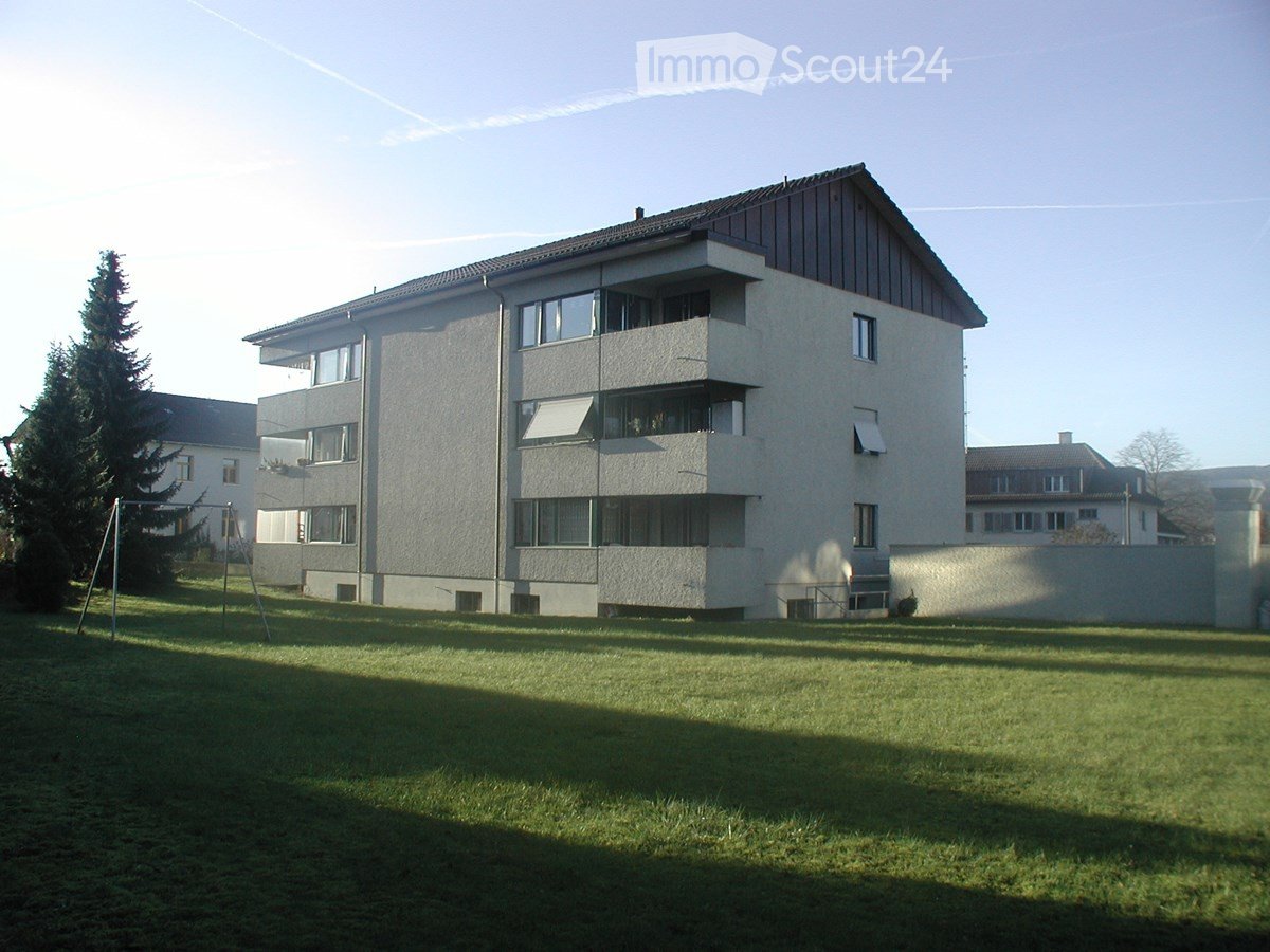 A multi-story residential building with balconies, surrounded by a grassy lawn and trees.
