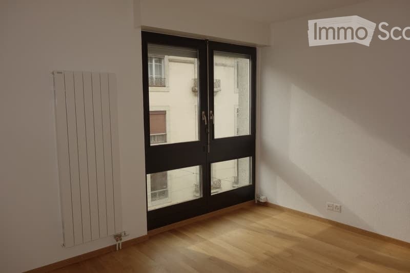 Empty room with wooden flooring, white walls, and large black-framed sliding doors leading to a balcony. A white radiator is mounted on the left wall.