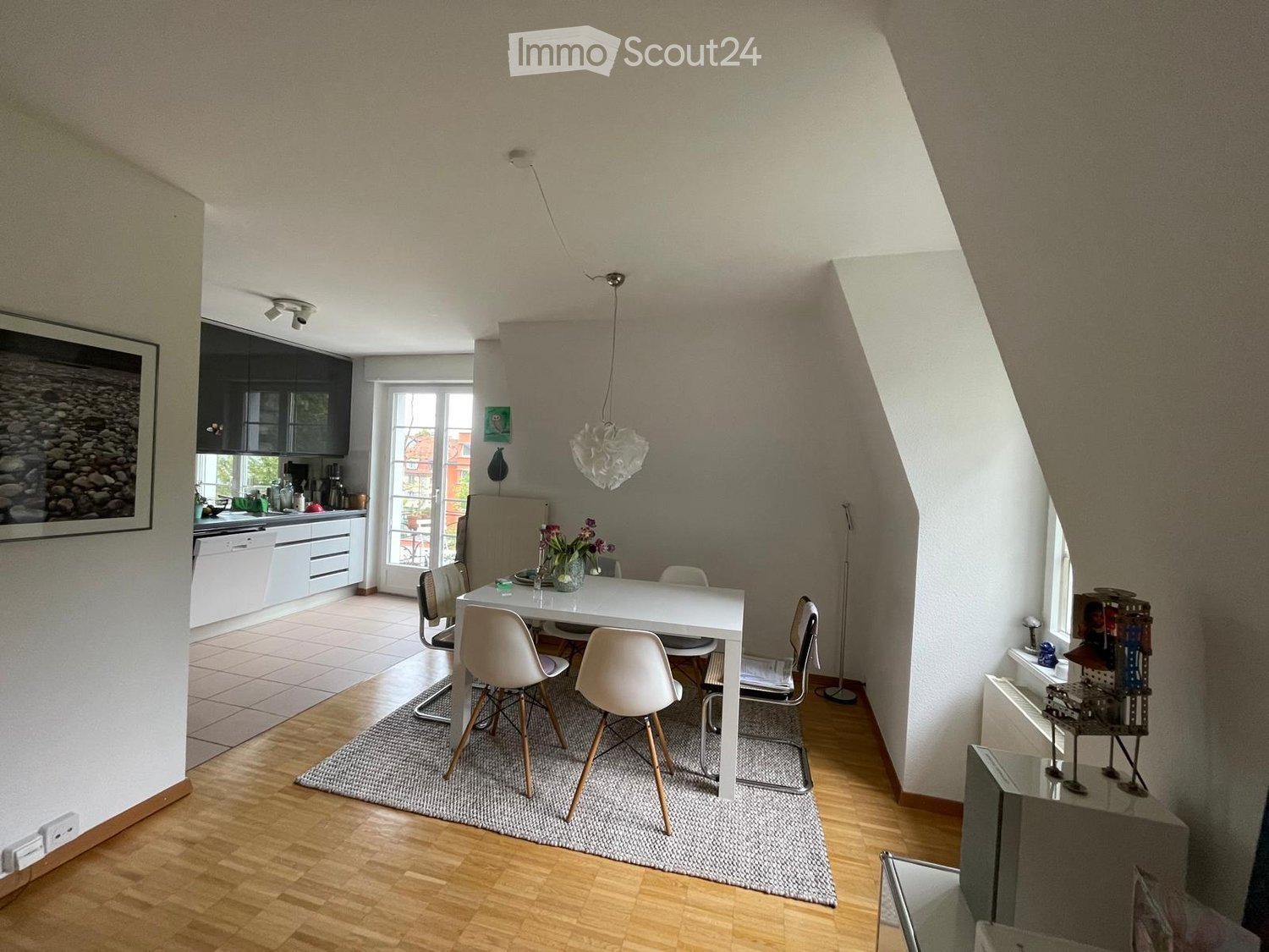 Bright, open-plan living and dining area with wooden flooring, white dining table, modern chairs, kitchen area with cabinets and appliances, and decorative items including a framed picture and a vase with flowers.