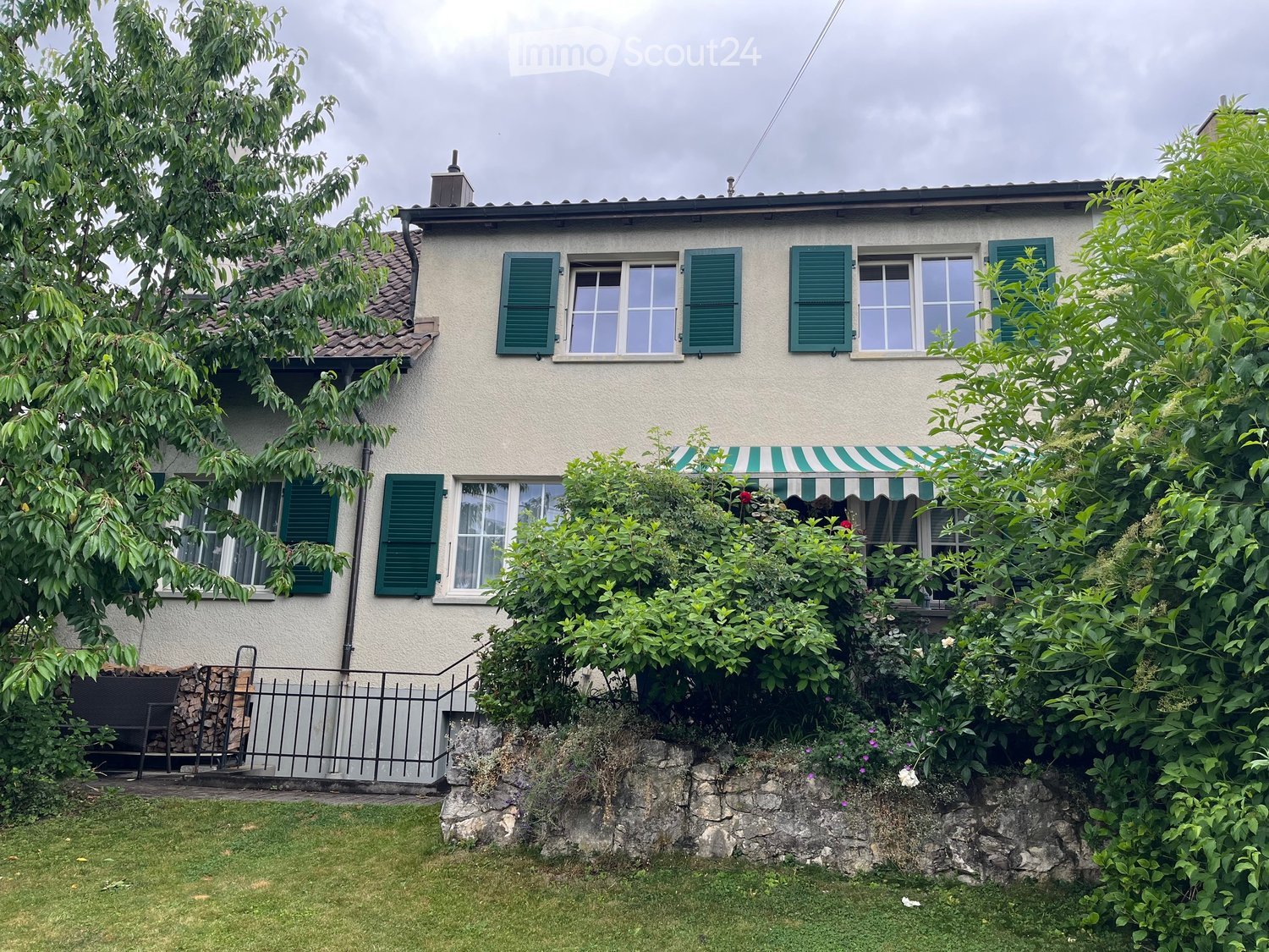 Two-story house with white walls, green shutters, and a tiled roof, surrounded by lush greenery and a stone retaining wall.