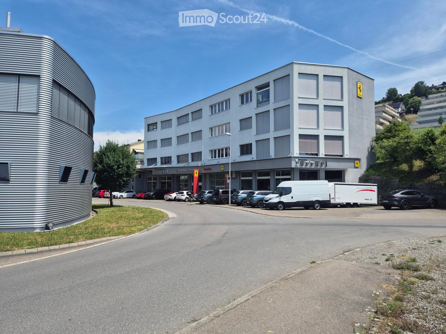 Modern building with large windows and a Ferrari logo, surrounded by parked cars and a paved area.