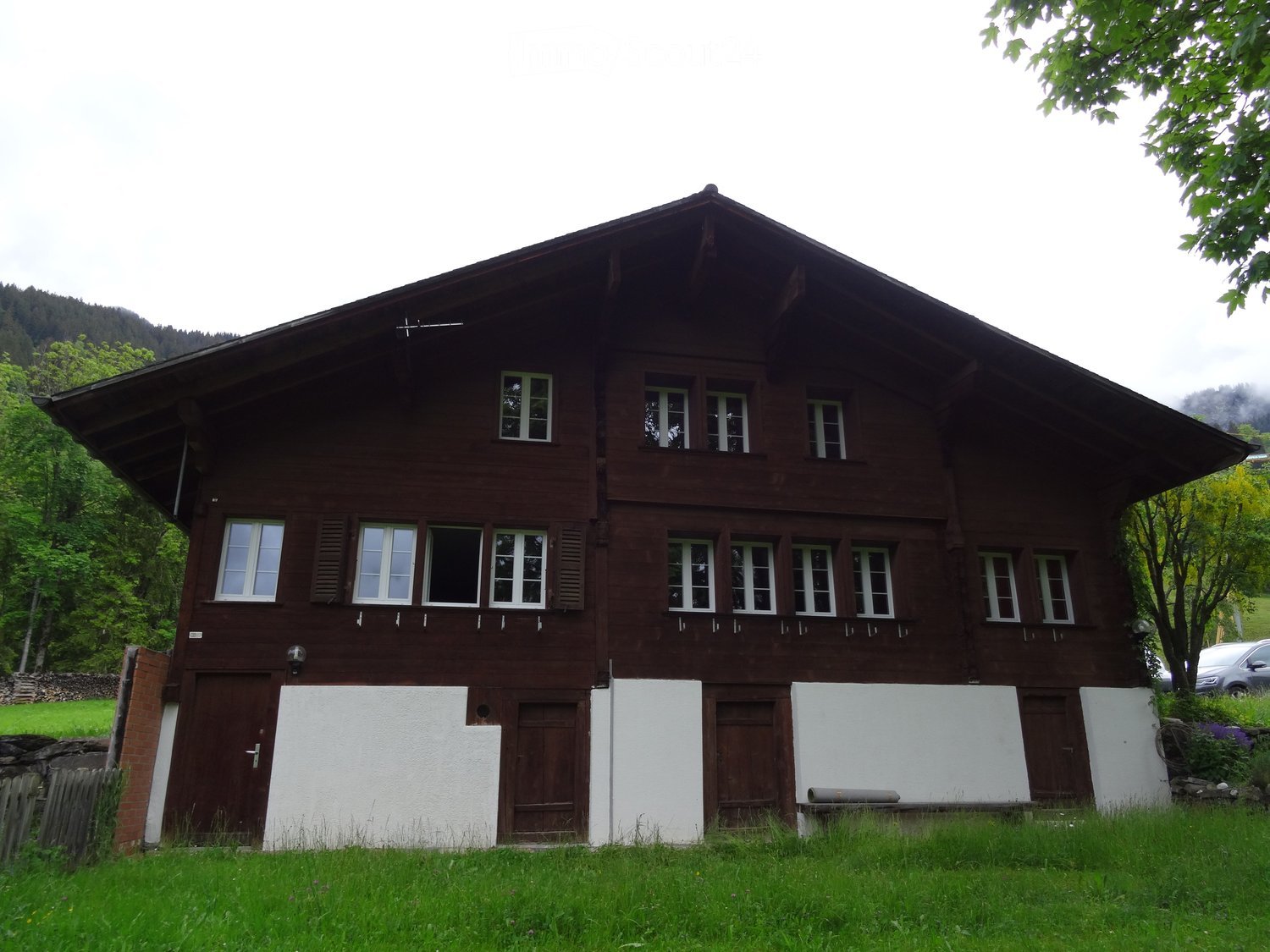 Traditional two-story wooden house with white trim, multiple windows, and a sloped roof, surrounded by greenery and a grassy lawn.