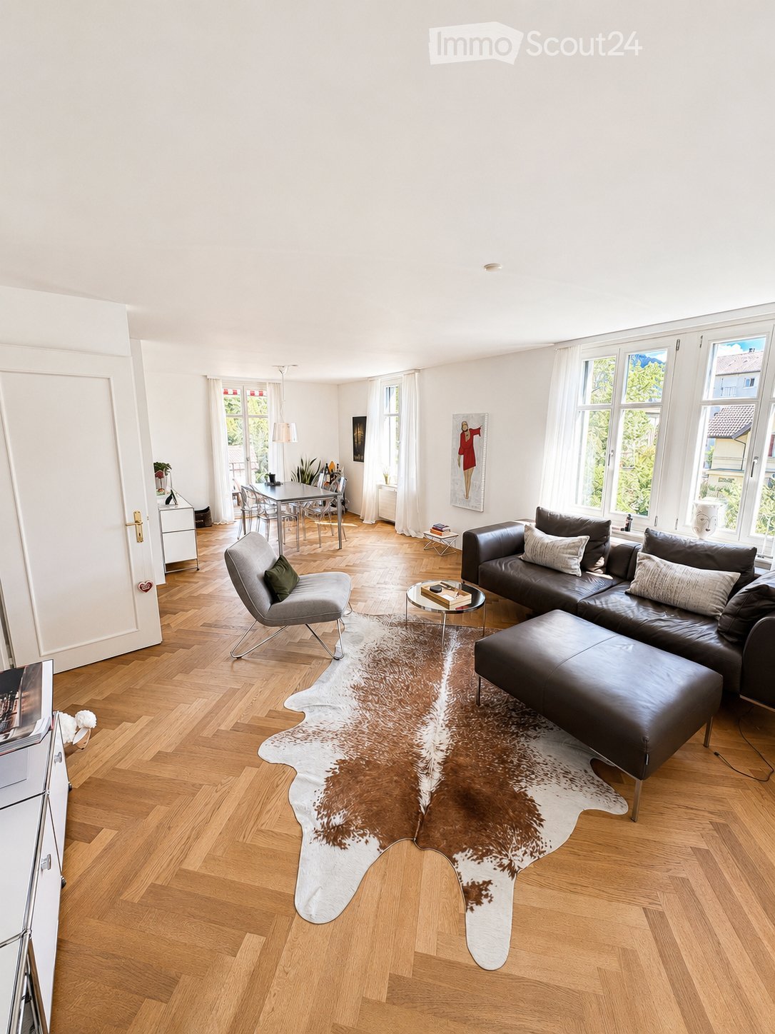 Bright living room with wooden flooring, large windows, a cowhide rug, a black leather sofa, an ottoman, a coffee table, a grey armchair, a dining area with a table and chairs, and a floor lamp.