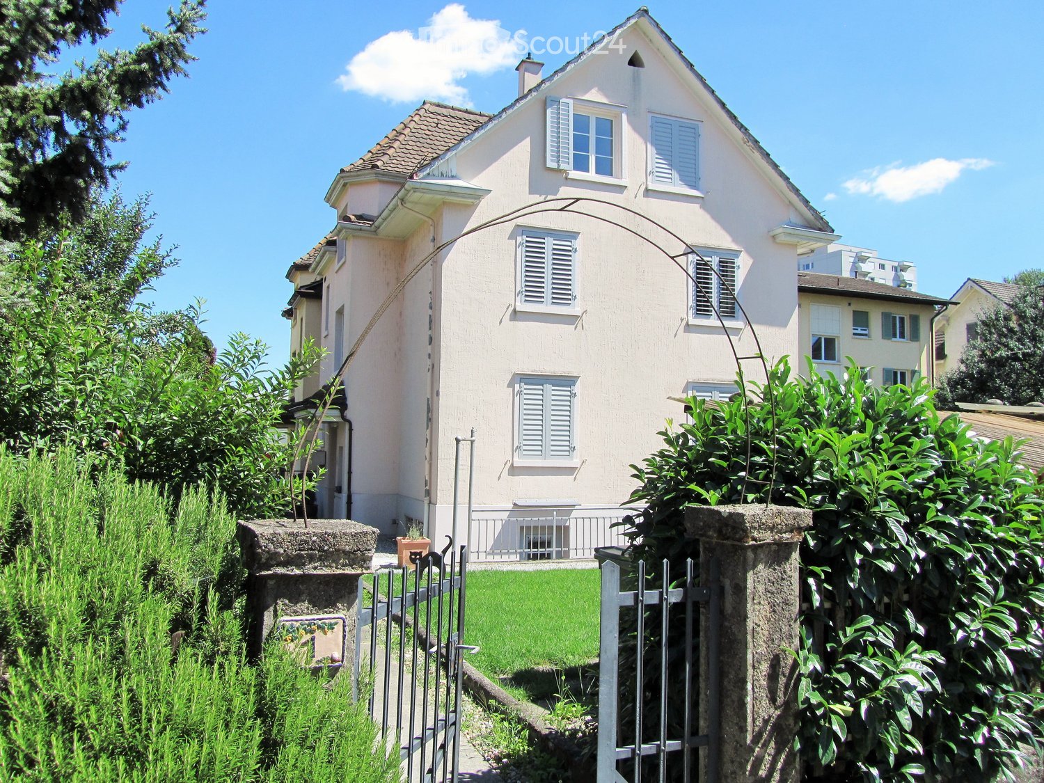 Two-story house with light pink exterior, multiple windows with shutters, and a well-maintained garden with a metal gate.
