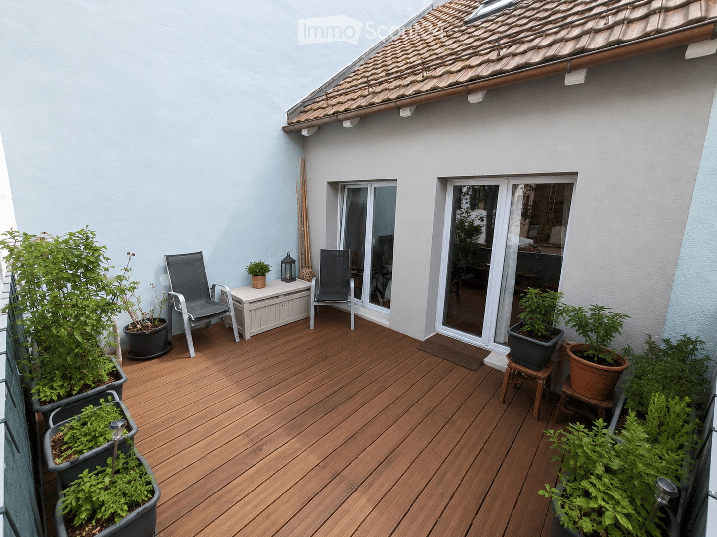 Wooden deck terrace with two chairs, a small table, potted plants, and a lantern. The terrace has a slanted roof and overlooks a building entrance.