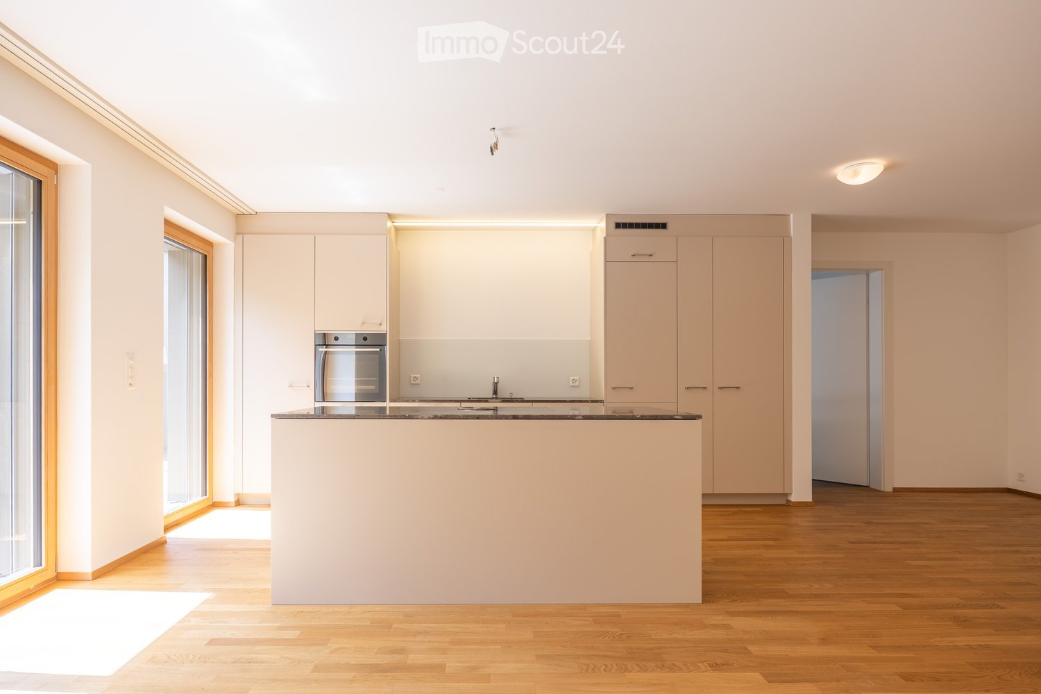 Modern kitchen with light-colored cabinets, a central island, and a built-in oven. The space features wooden flooring and large windows allowing natural light to fill the room.