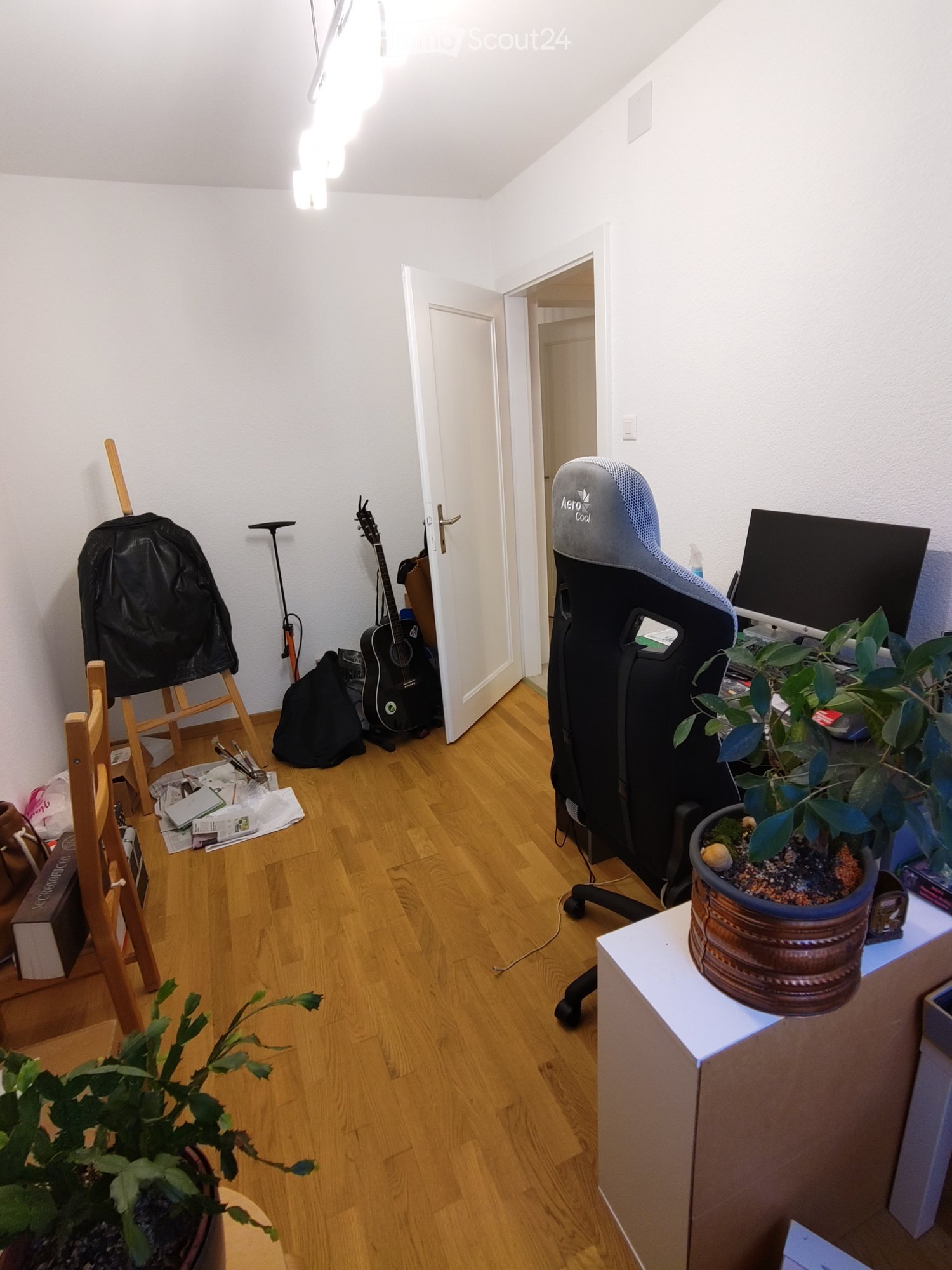 A furnished room with wooden flooring, a chair, a guitar, a backpack, a screen, and plants.