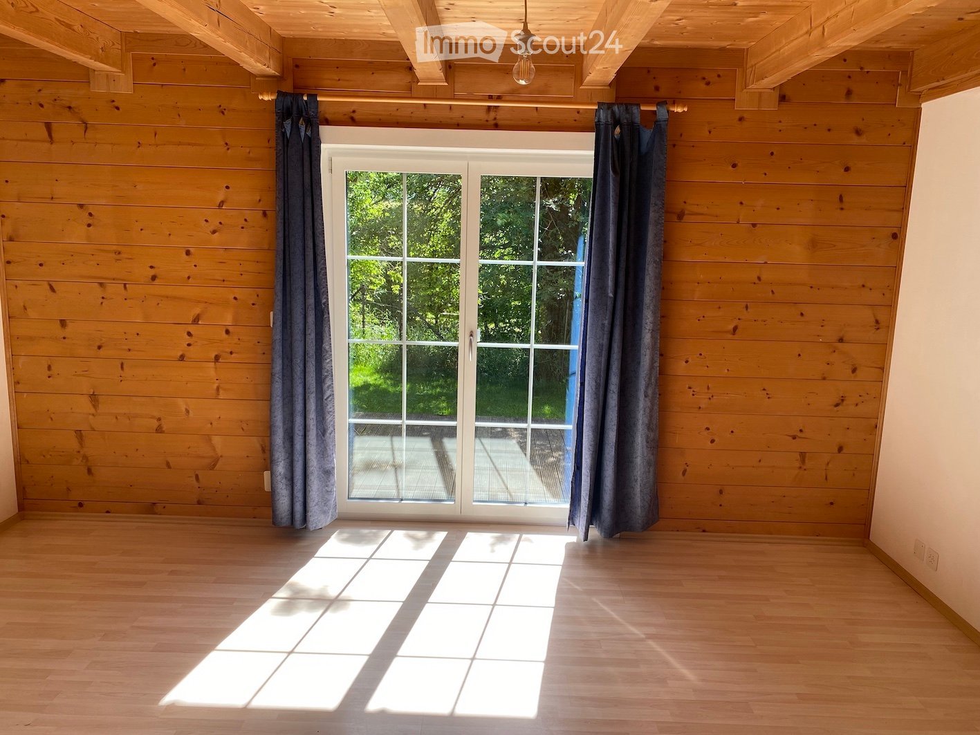 Empty room with wooden walls and floor, large sliding glass door with curtains, natural light streaming in.