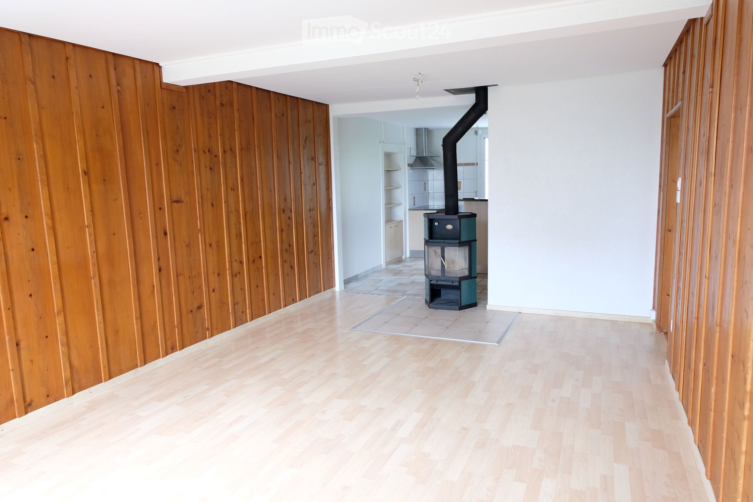 Empty room with light wooden flooring and paneled wooden walls, featuring a wood stove in the center and a view into a kitchen area with white cabinets and a stainless steel range hood.