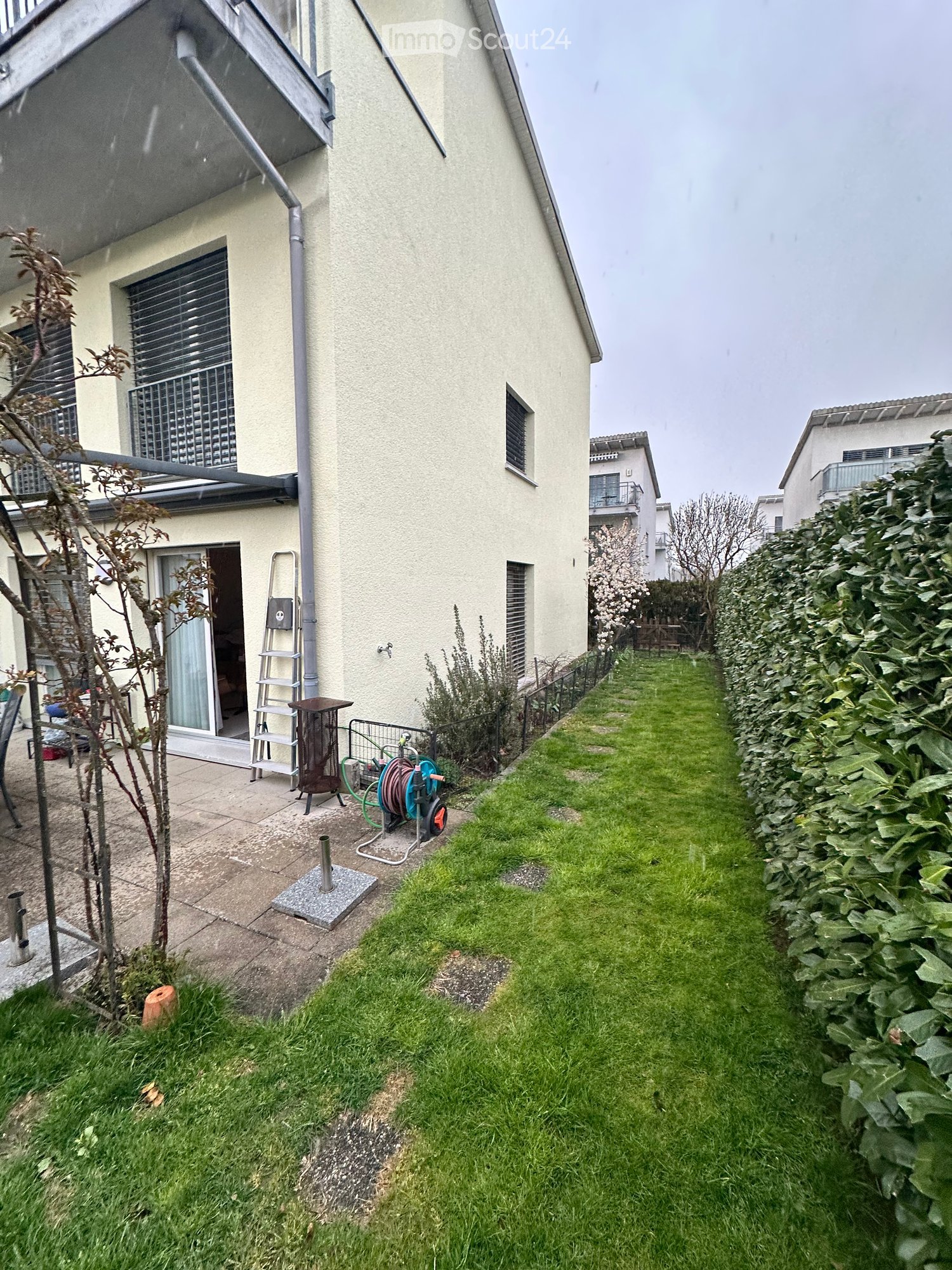 Garden area with a well-maintained lawn, a small patio with a ladder, a hose reel, and a small tree. The house has a light-colored exterior with a covered entrance and a window with blinds.